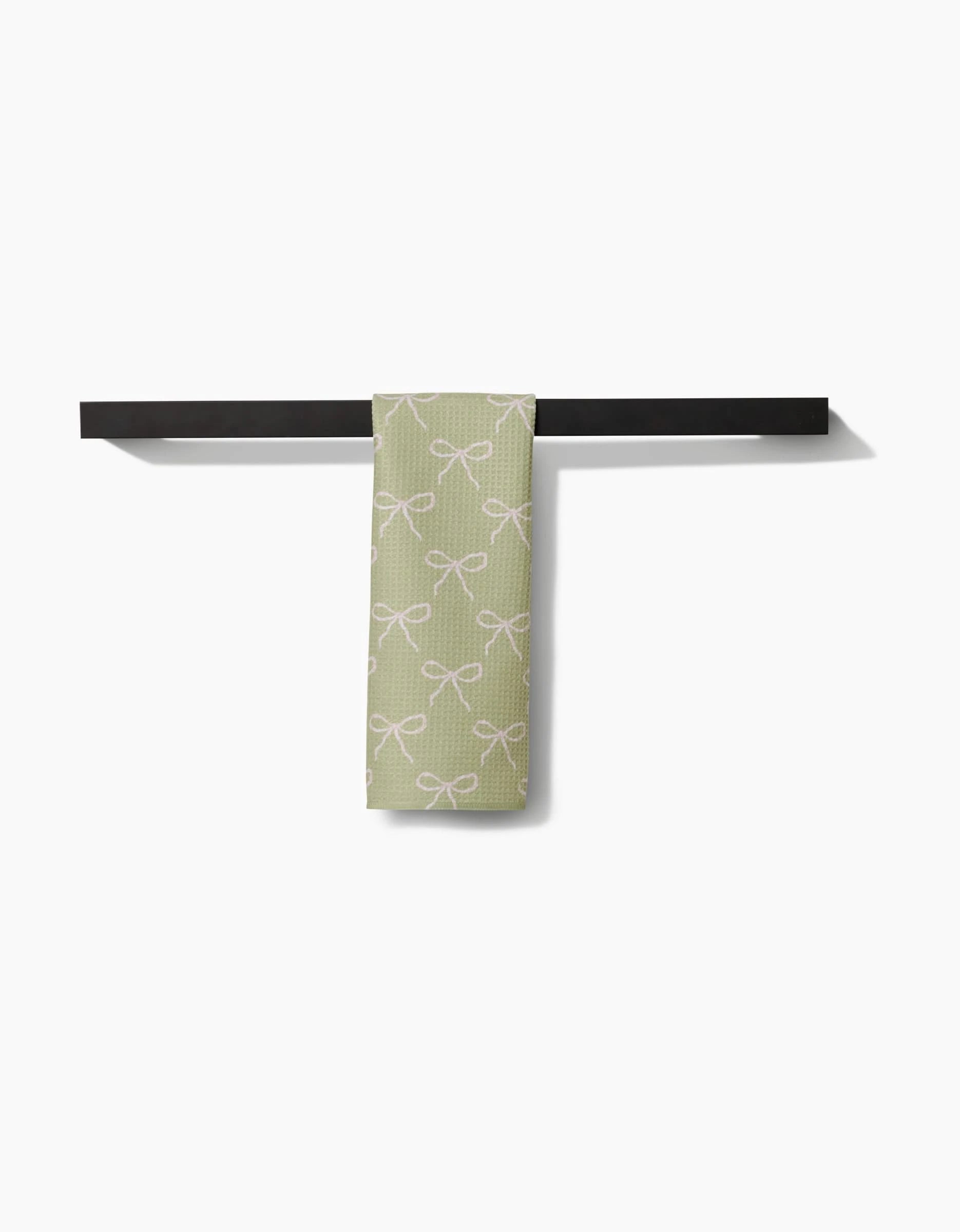 The Laura Prietto Lattice Bows Bar Towel, featuring a white bow pattern on light green ultra-absorbent Geoweave fabric, is draped over a modern black towel bar set against a plain white background.
