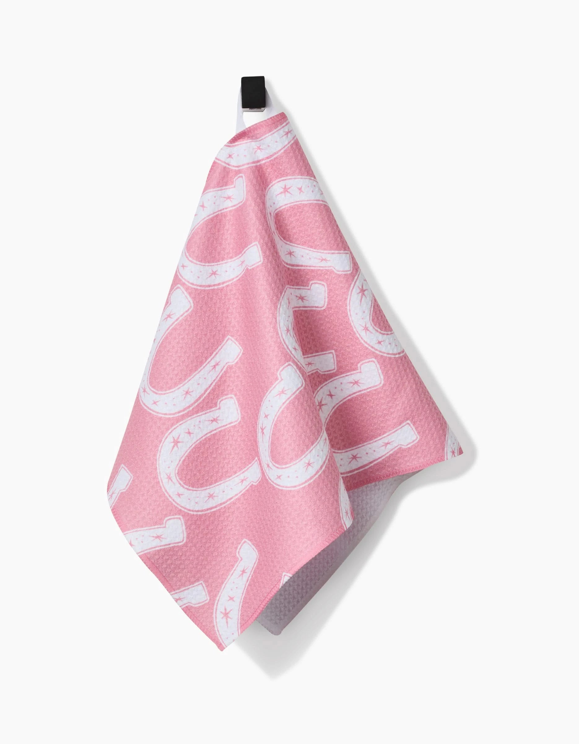 The Lucky Horseshoe Bar Towel by Aubrey Fairchild, made from Geoweave fabric, features a pink design with white horseshoes and small stars. It hangs from a black loop against a white background.