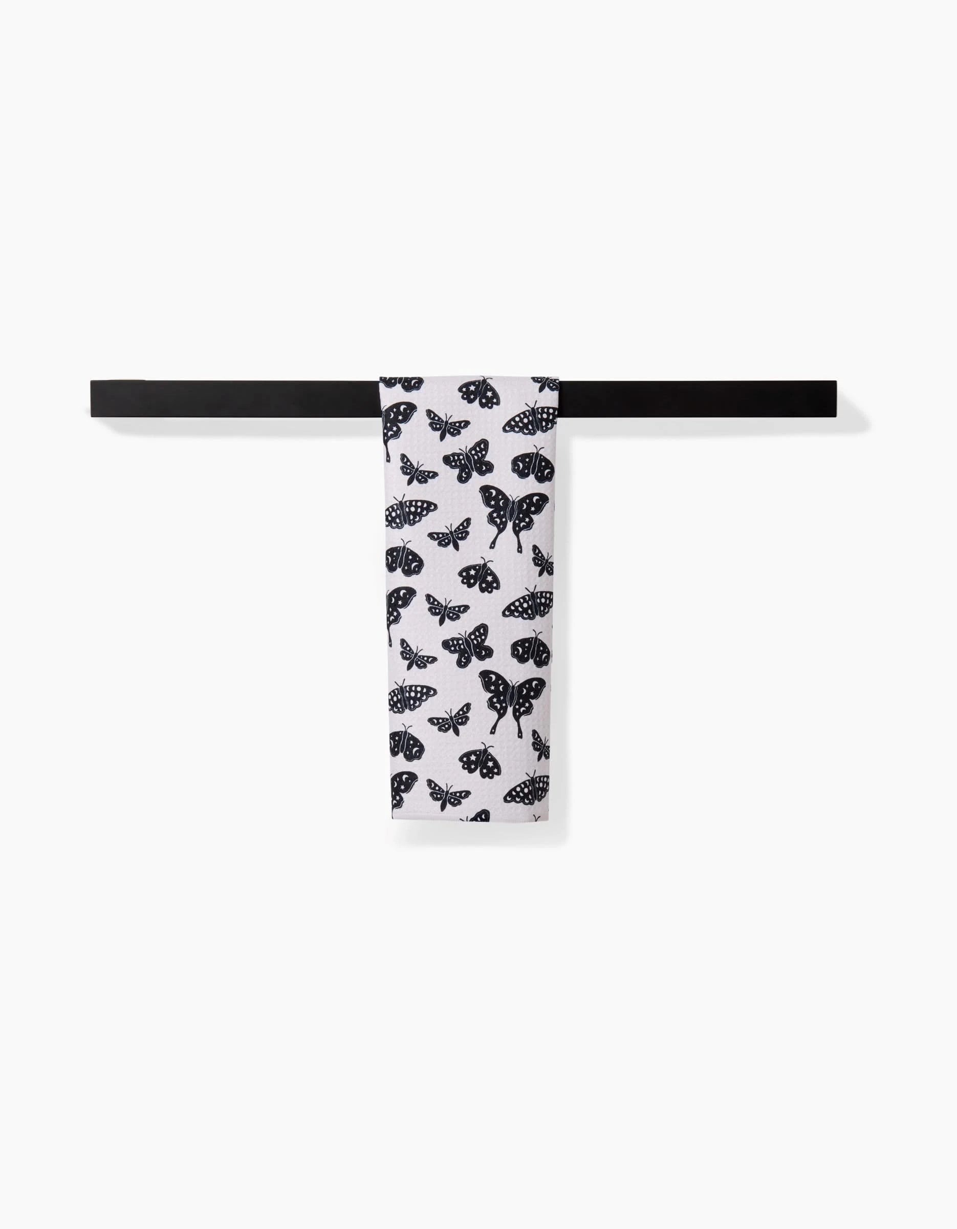 The Kayla Ann Lunar Moths White towel, an ultra-absorbent white towel with a black moth design, hangs neatly over a horizontal black bar against a plain white background.