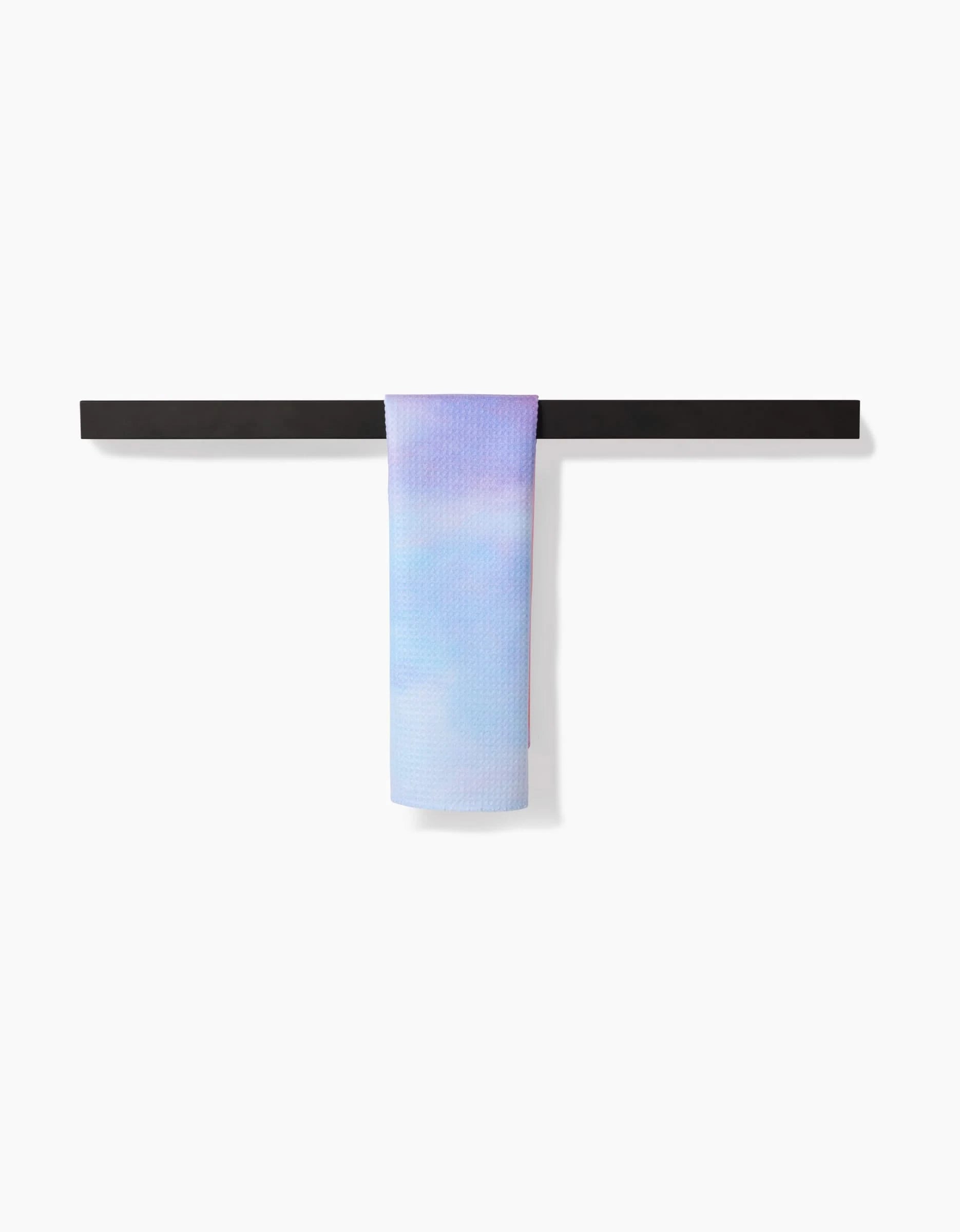 The Magic In The Sky Bar Towel by Stefanie Bales, featuring pastel blue and lavender Geoweave™ fabric, is draped over a minimalist black towel rack against a plain white background.