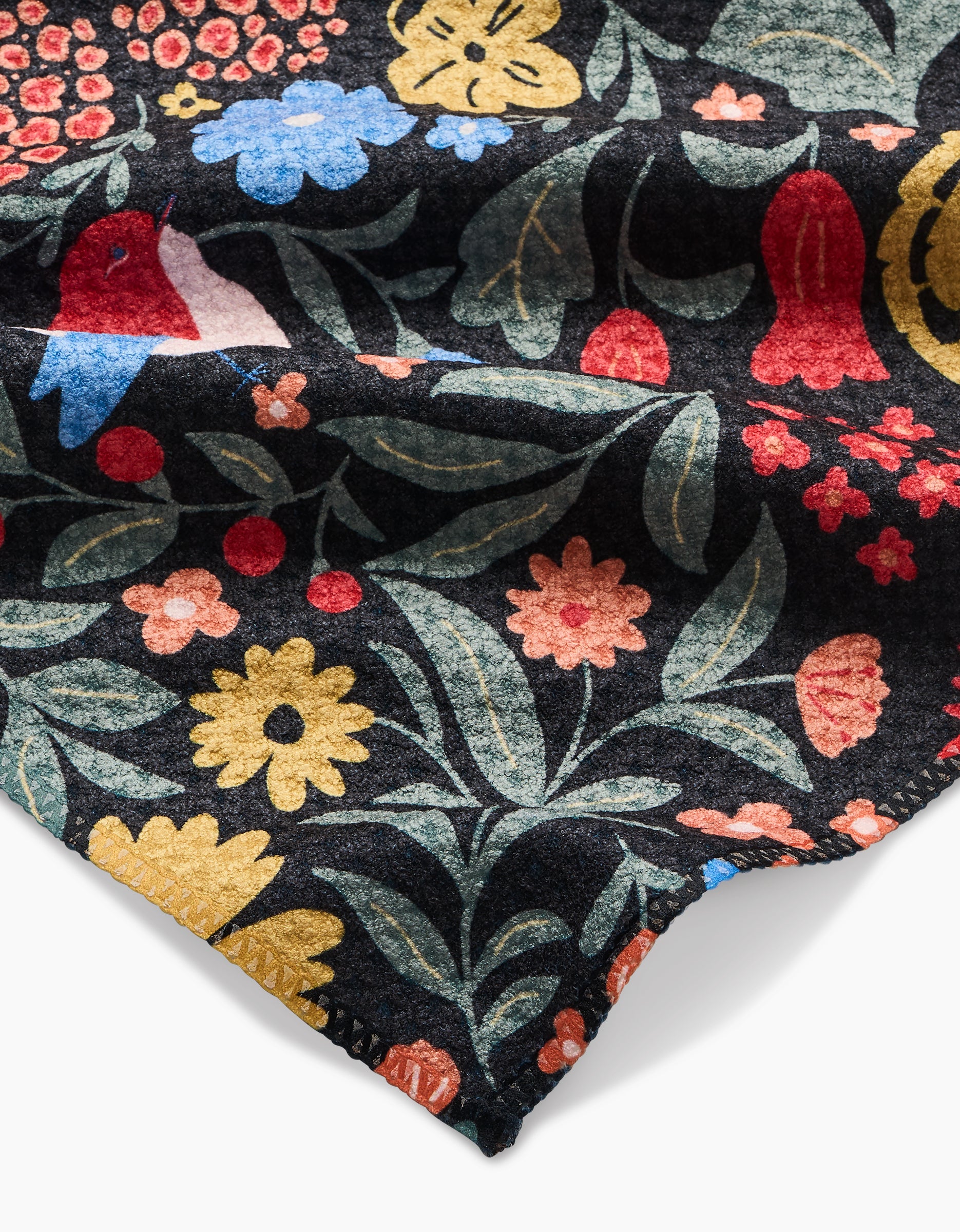 A close-up of Danelys Sidron’s Marsh Wren bar towel shows ultra-absorbent Geoweave fabric with a colorful floral and bird pattern on dark background—yellow, red, blue, and pink flowers with green leaves decorate this stylish towel.