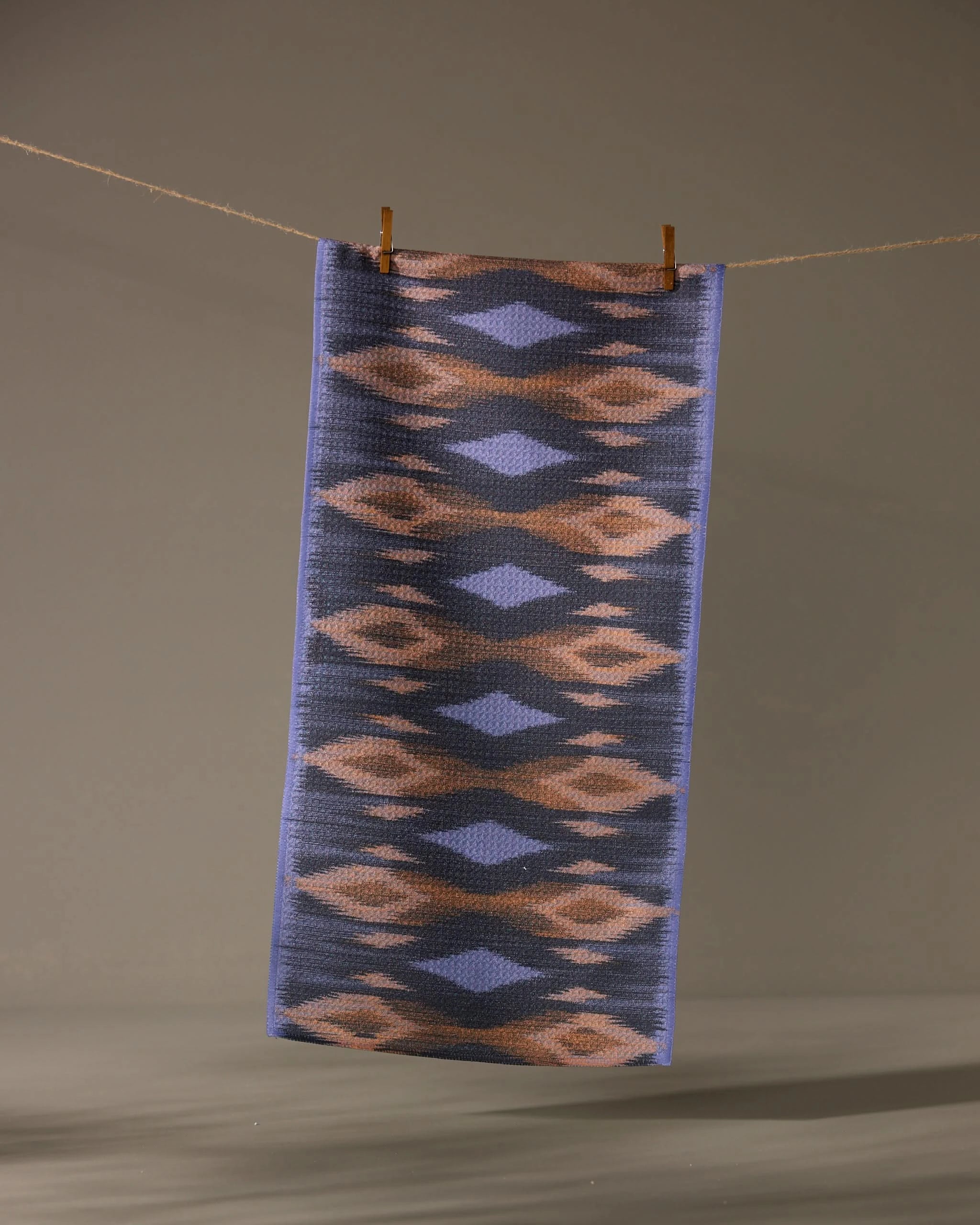 The Geometry Mesa towel, a rectangular ultra-absorbent piece with a blue, brown, and beige geometric pattern, hangs from a clothesline against a plain background, secured by two wooden clothespins.