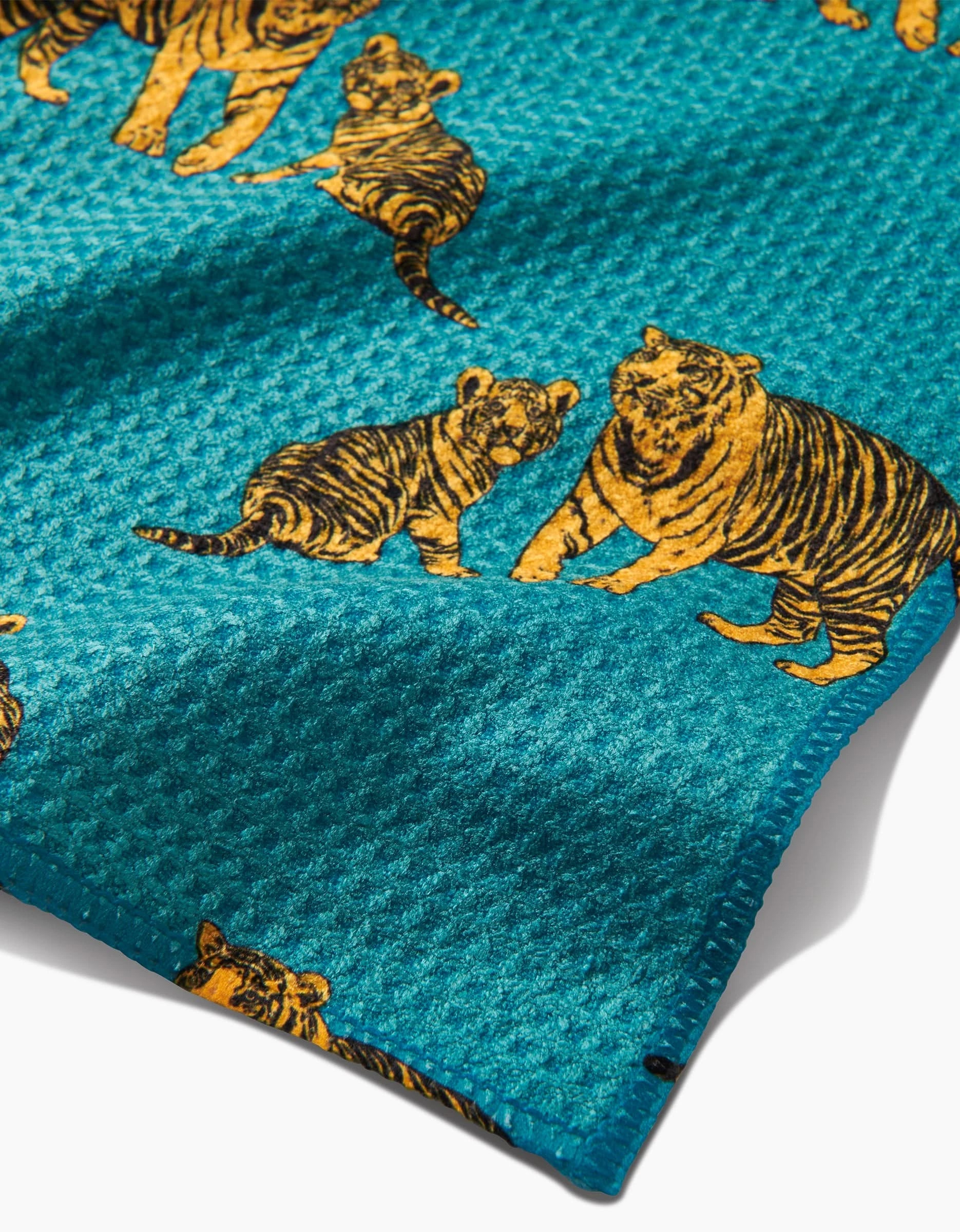 A close-up of the Sarah Holden Mommy and Me Bar Towel, made from textured teal Geoweave fabric, featuring a repeating pattern of illustrated orange and black tigers with their cubs.