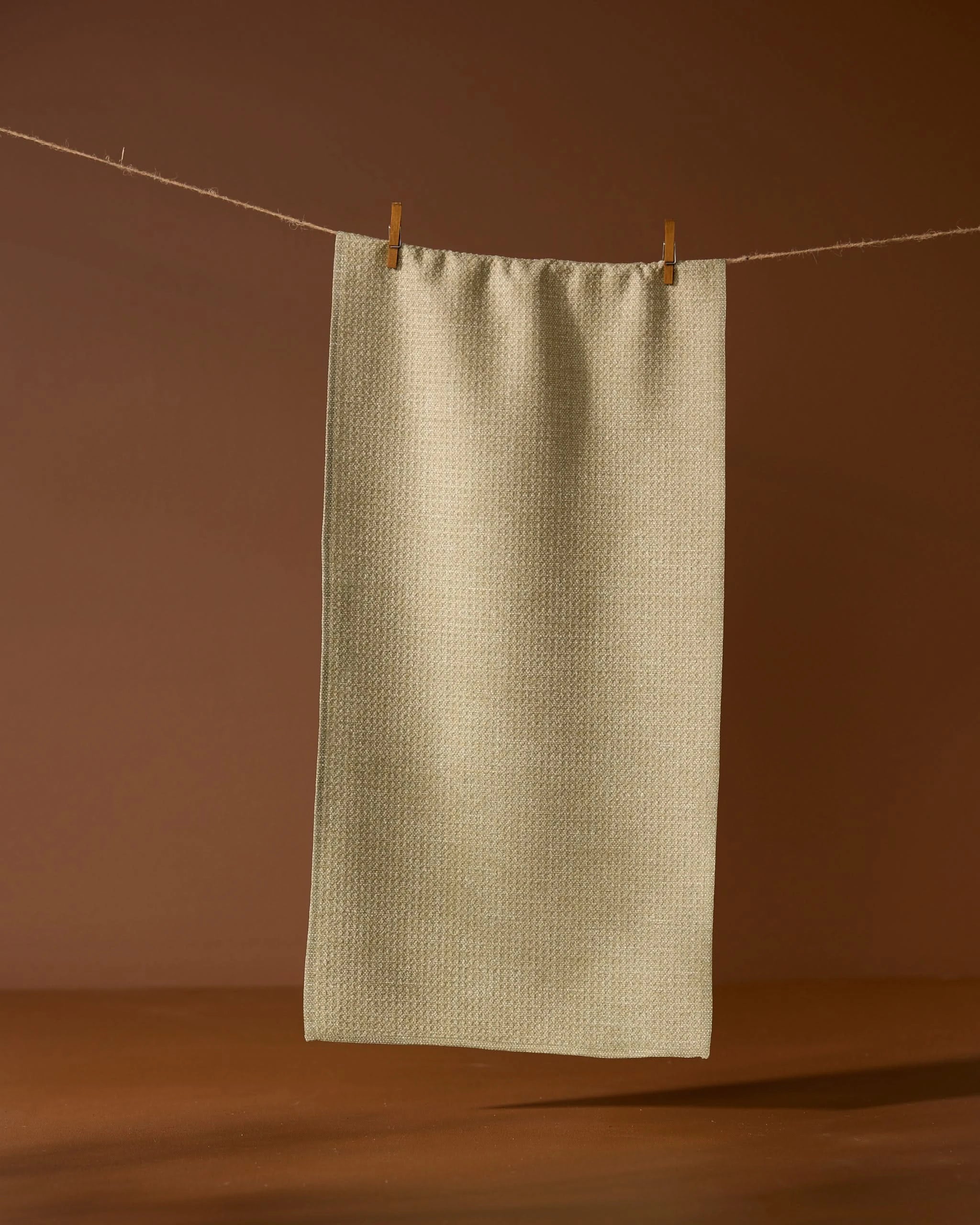 The Geometry Mosswood recycled kitchen towel in light beige hangs from a string with two wooden clothespins, set against a brown background.