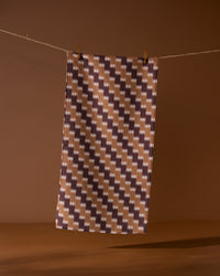 The Geometry Mountain Song bar towel features a brown, beige, and dark purple houndstooth pattern and hangs against a brown background. Made from recycled materials, it’s an eco-friendly and stylish choice.