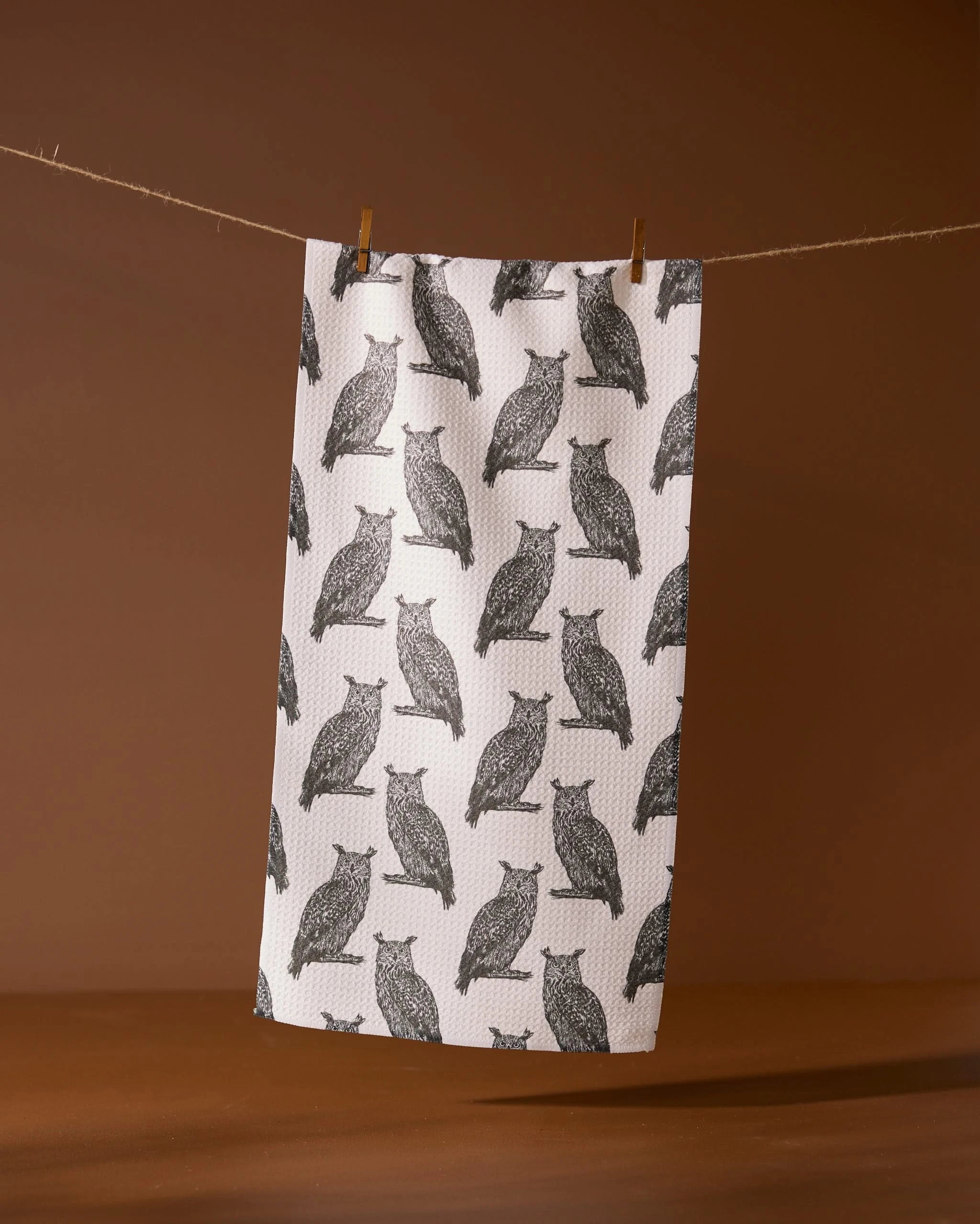 The Geometry Nocturne towel, featuring a black owl pattern on white, hangs on a clothesline with two wooden clothespins against a brown background.