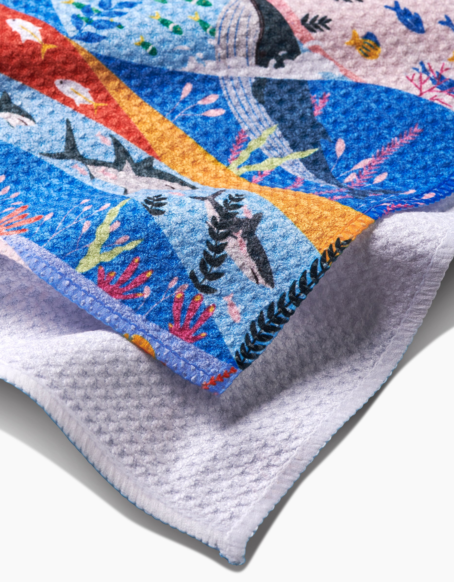 The Oceans towel by Ceyda Alasar, made from recycled materials and decorated with vibrant sharks, dolphins, seaweed, and other marine life, is folded to show its textured white underside.