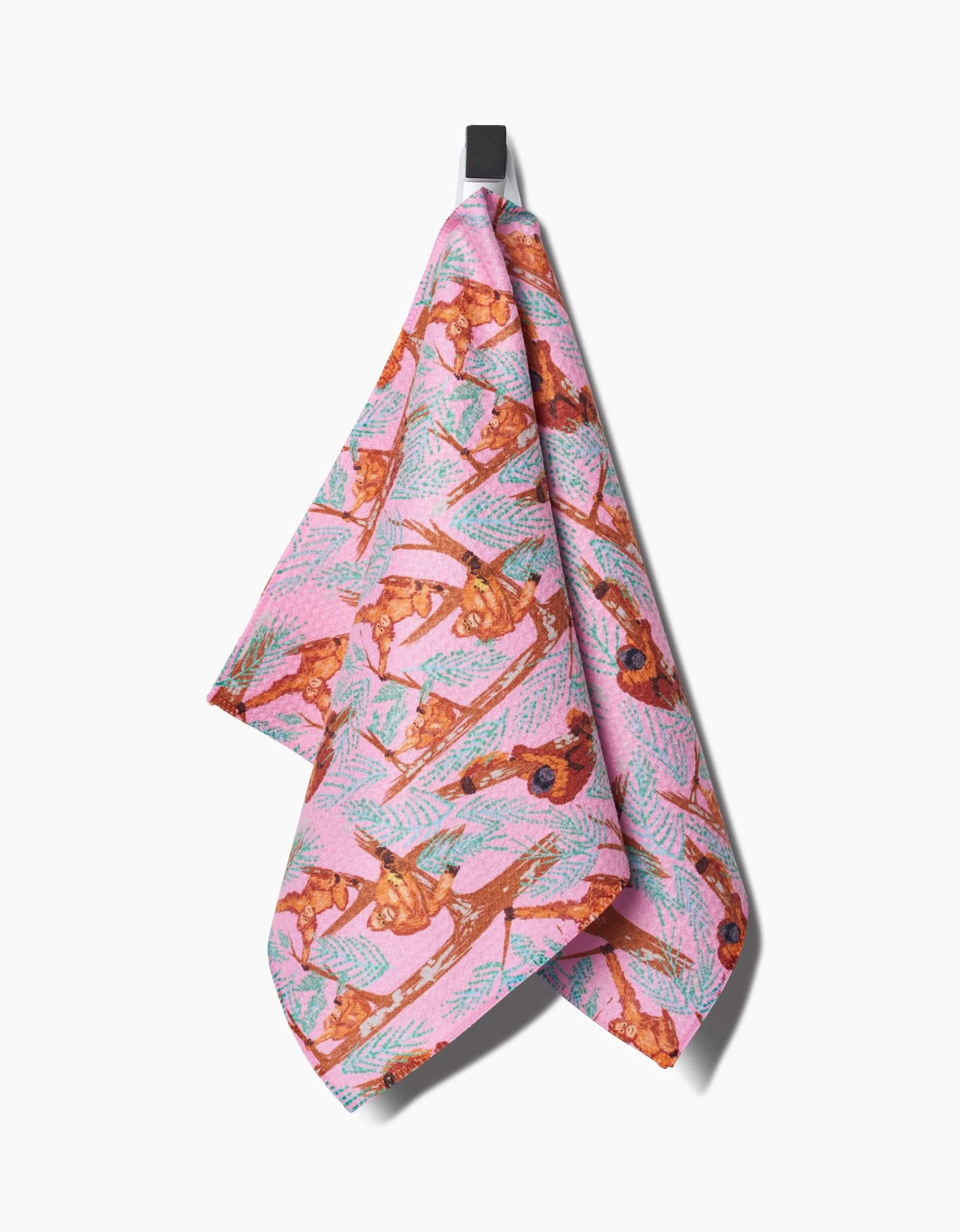 The Orangutan Clan bar towel by Elisabeth Wing, made from recycled materials, showcases brown squirrels and green pine branches on pink fabric and hangs from a black hook against a white background.