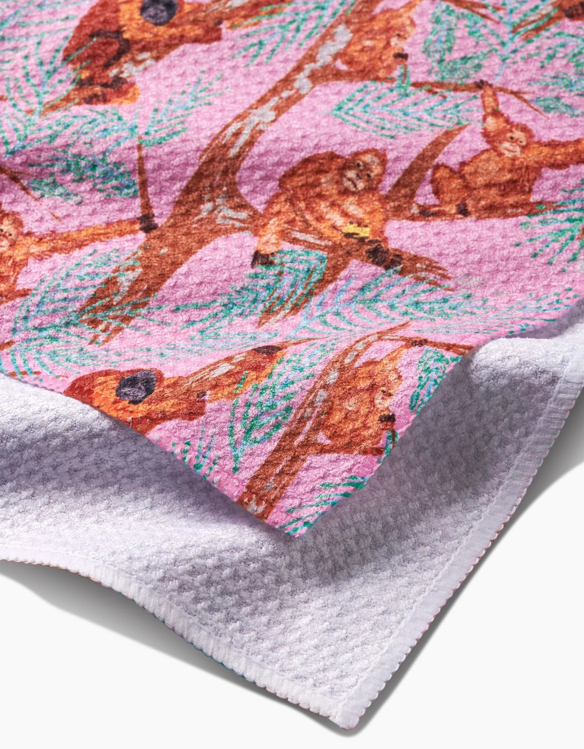A close-up of the Orangutan Clan bar towel by Elisabeth Wing, set against a pink background. It shows brown orangutans on tree branches with green leaves, has a textured white underside, stitched edges, and is made from recycled materials.