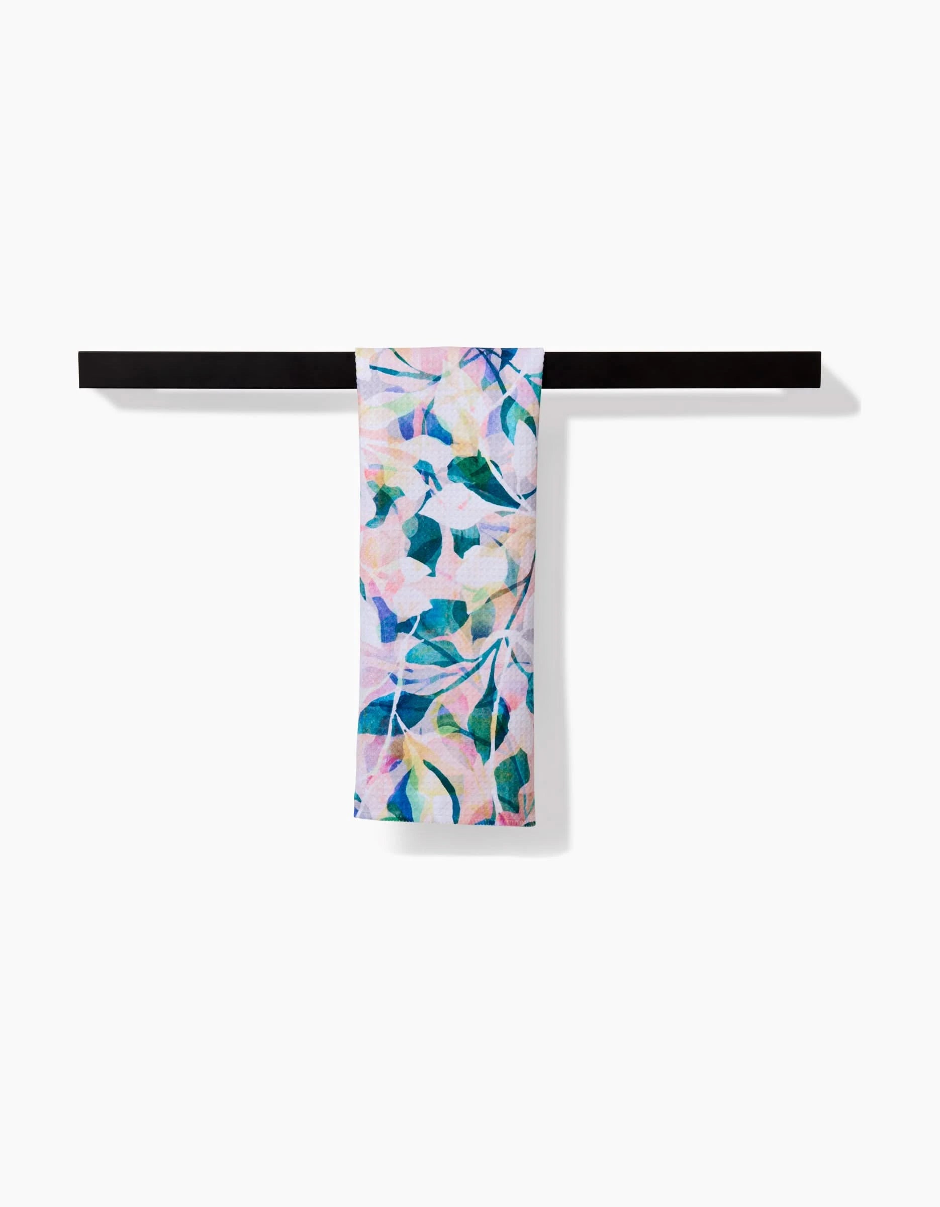 The Rachel Parker Painted Foliage bar towel, featuring a vibrant botanical design and made from recycled materials, is displayed draped over a sleek black bar against a white background.
