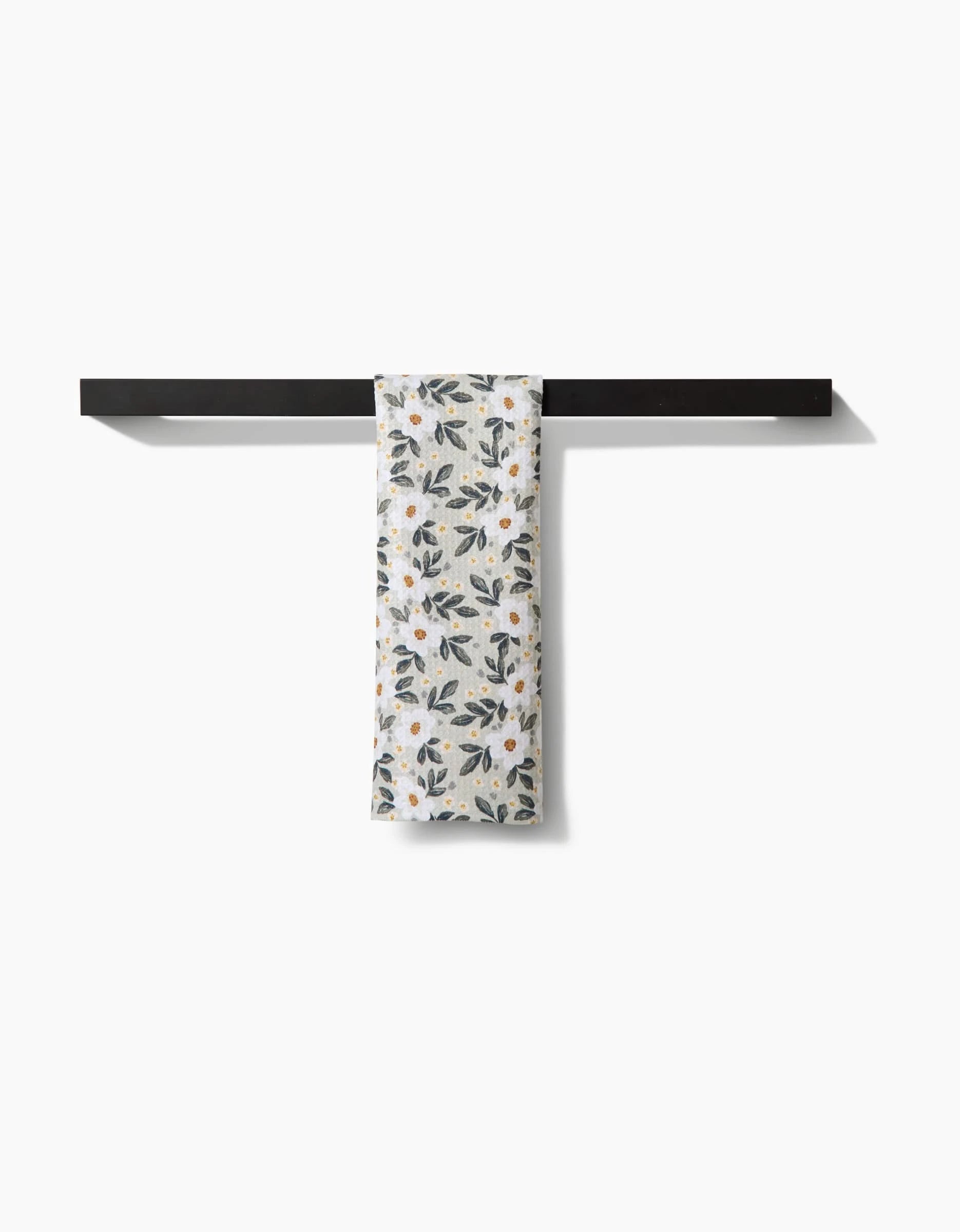 A white Painted Spring Flowers bar towel by Teri the Butter, adorned with blue and yellow blooms, is neatly draped over a sleek black towel bar against a plain white background.