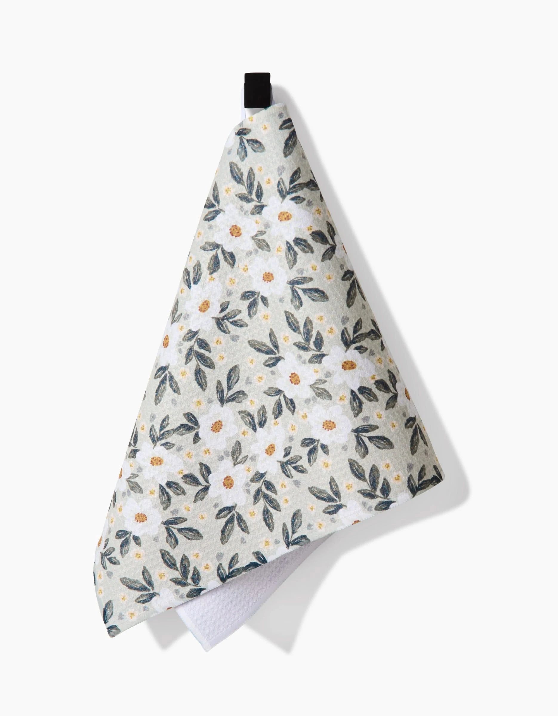 The Teri the Butter Painted Spring Flowers Bar Towel, featuring green leaves and white flowers on Geoweave fabric, hangs from a black loop against a white background.