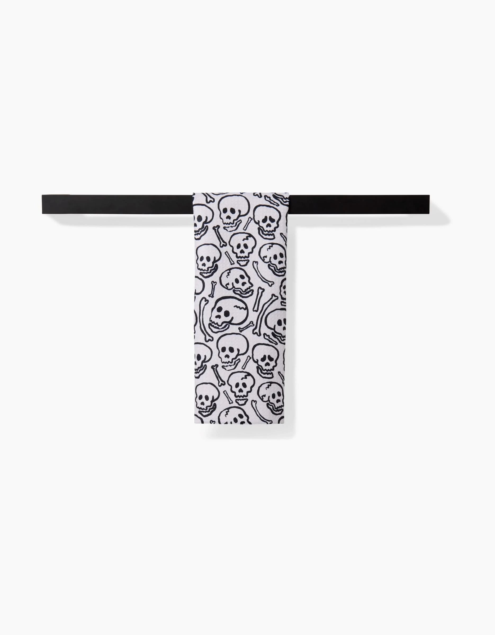 The Kayla Ann Painterly Skulls white ultra-absorbent towel, featuring a black skull and bones pattern, hangs neatly over a simple black towel bar against a plain white background.