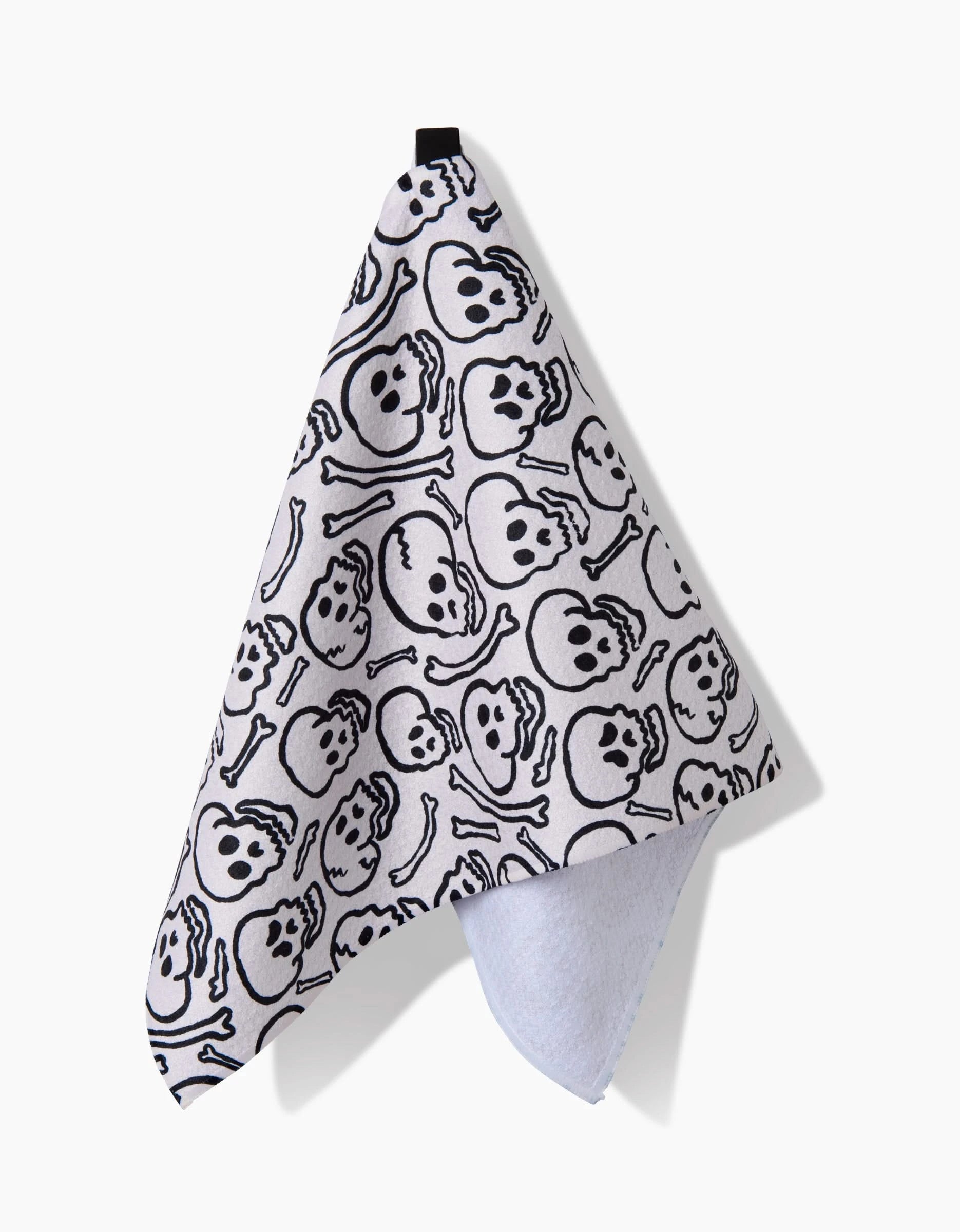 The Kayla Ann Painterly Skulls bar towel features black cartoon skull and crossbones on white, with a folded corner showing the plain underside. Hanging from a black loop, it’s made from recycled materials for an eco-friendly touch.