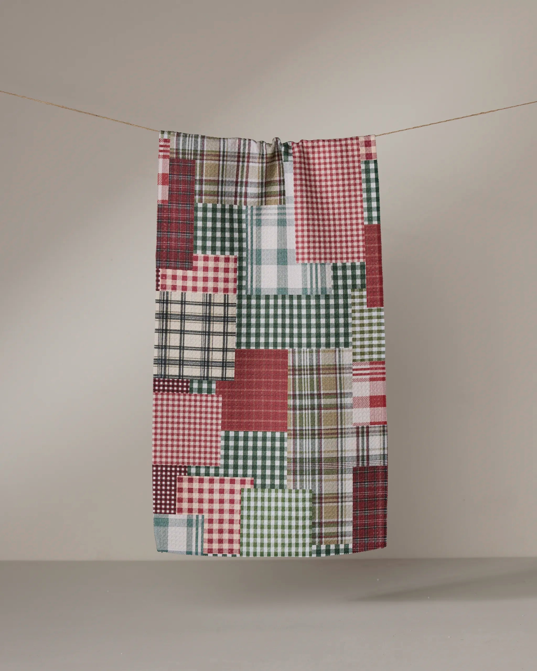 The Geometry Patchly bar towel, made from recycled materials, features red, green, white, and beige plaid and checkered patterns and hangs on a clothesline against a neutral grey background.