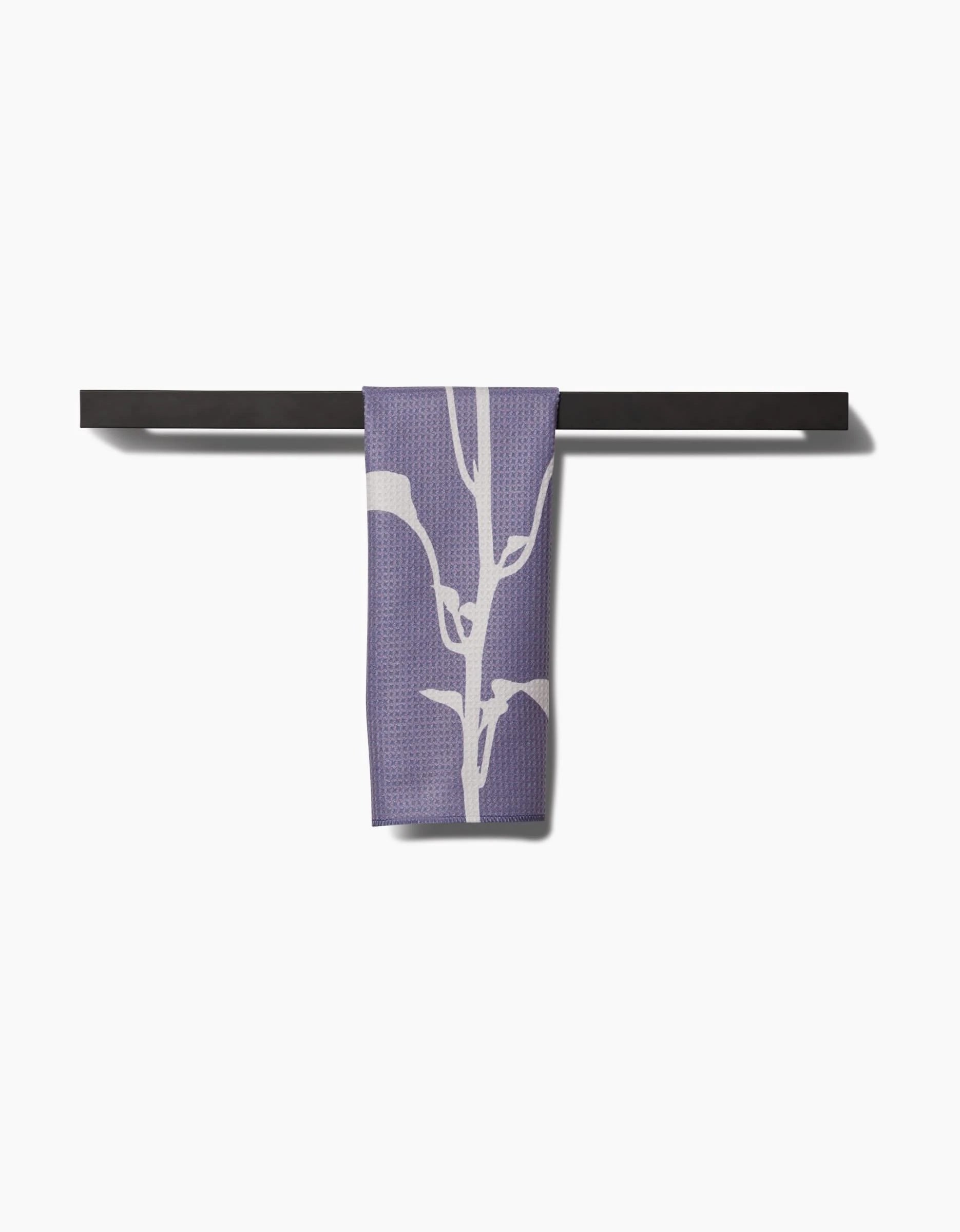 A PrintedPlant Pepper Bar Towel in purple with a white botanical design, made from Geoweave fabric, hangs over a black bar set against a plain white background.