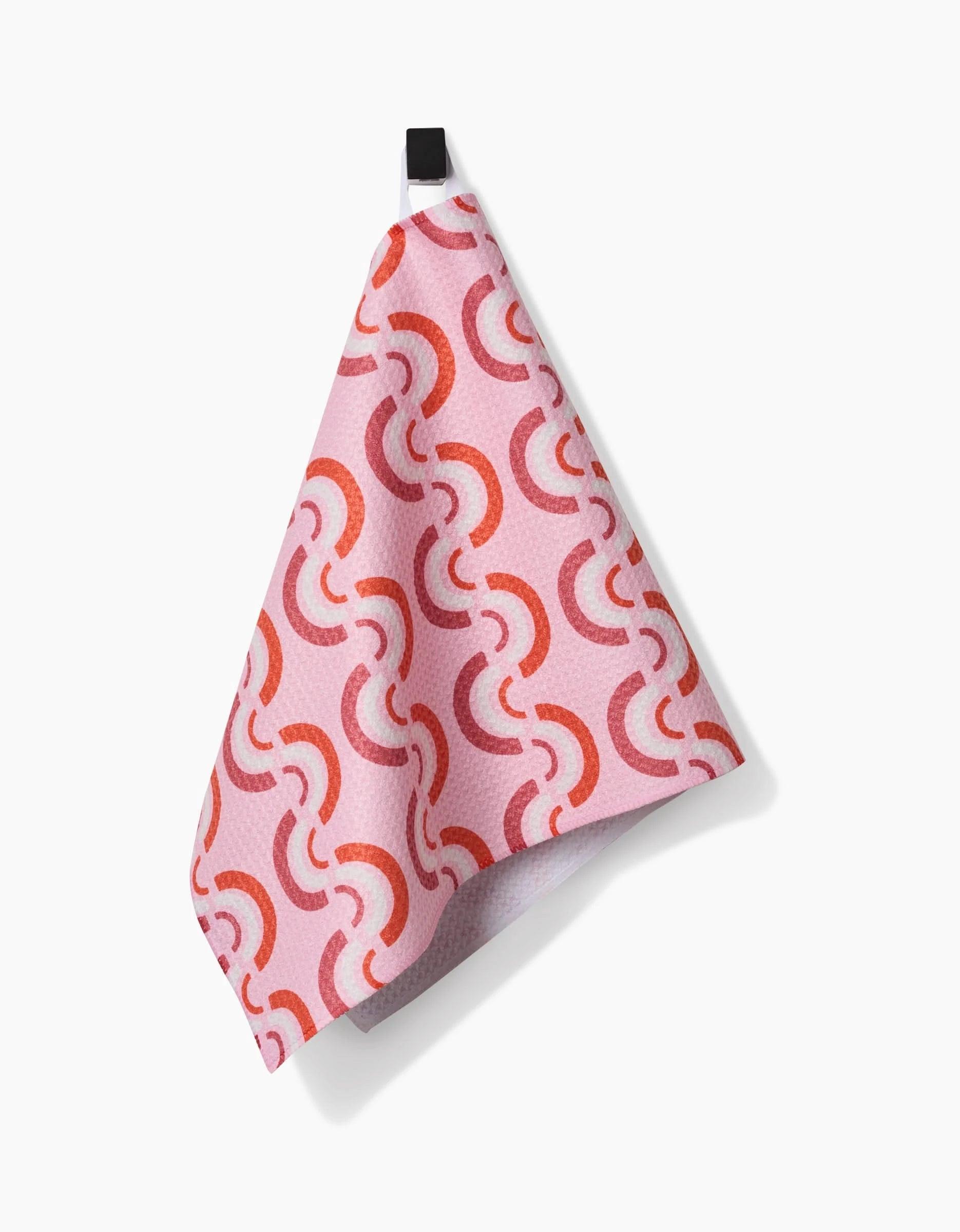 The Rainbow Waves bar towel by Whitney Urquhart, made from recycled materials with a red and orange curved geometric pattern, hangs from a black hook against a white background.