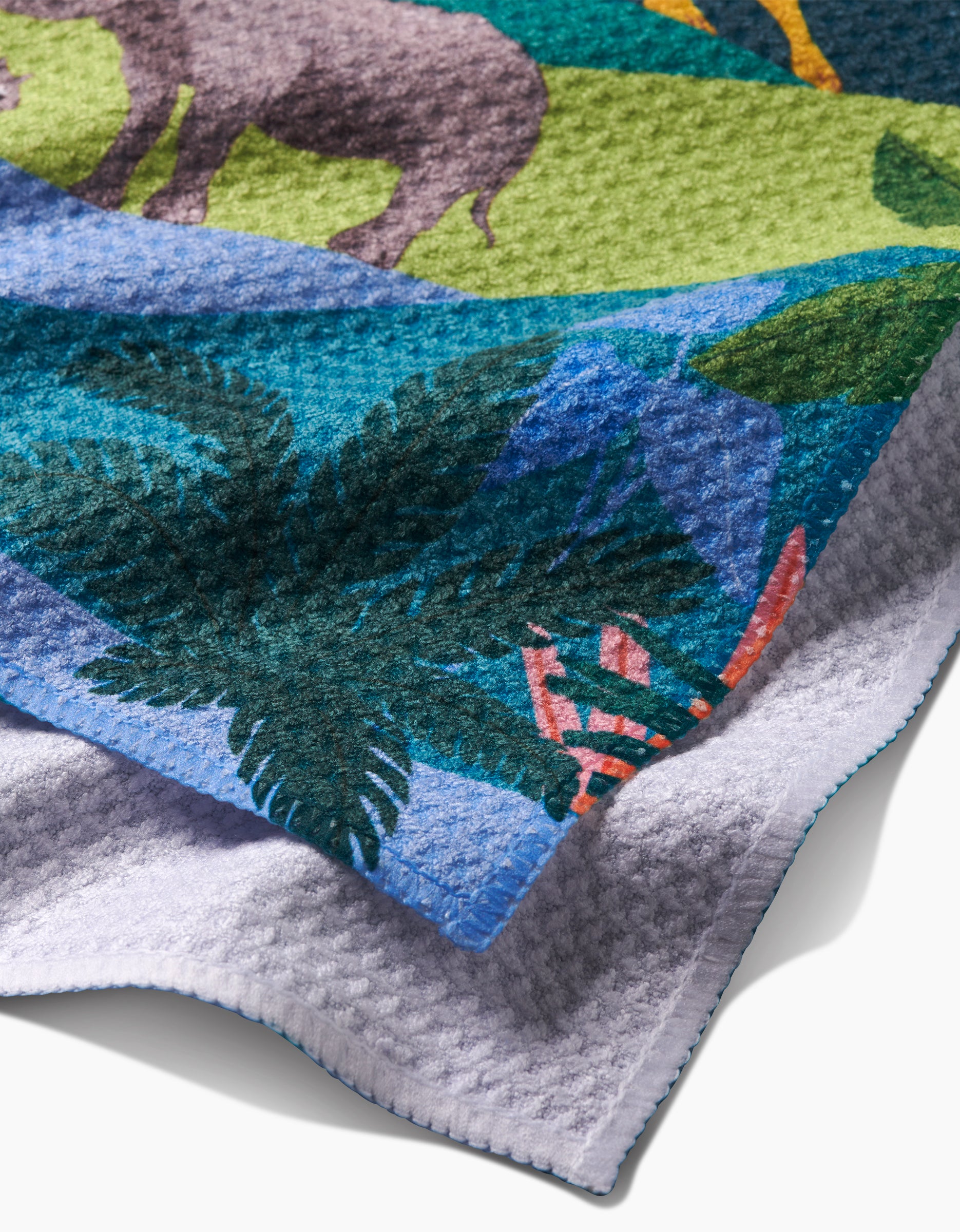A close-up of Ceyda Alasar’s “Rainforest” bar towel, made from recycled materials, shows its colorful safari design with a rhinoceros, green leaves, tropical plants, and its light gray underside.