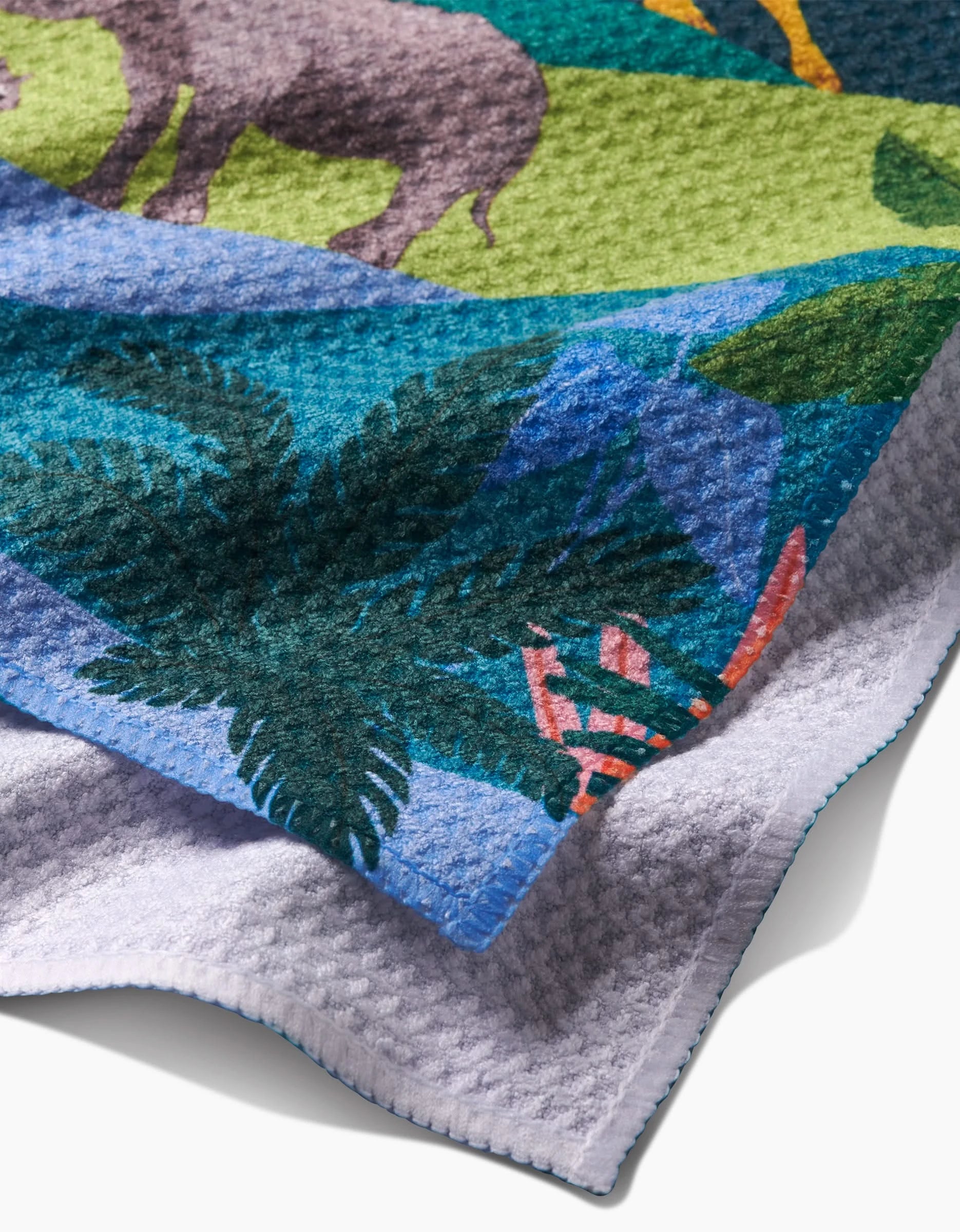 A close-up of Ceyda Alasar’s “Rainforest” bar towel, made from recycled materials, shows its colorful safari design with a rhinoceros, green leaves, tropical plants, and its light gray underside.