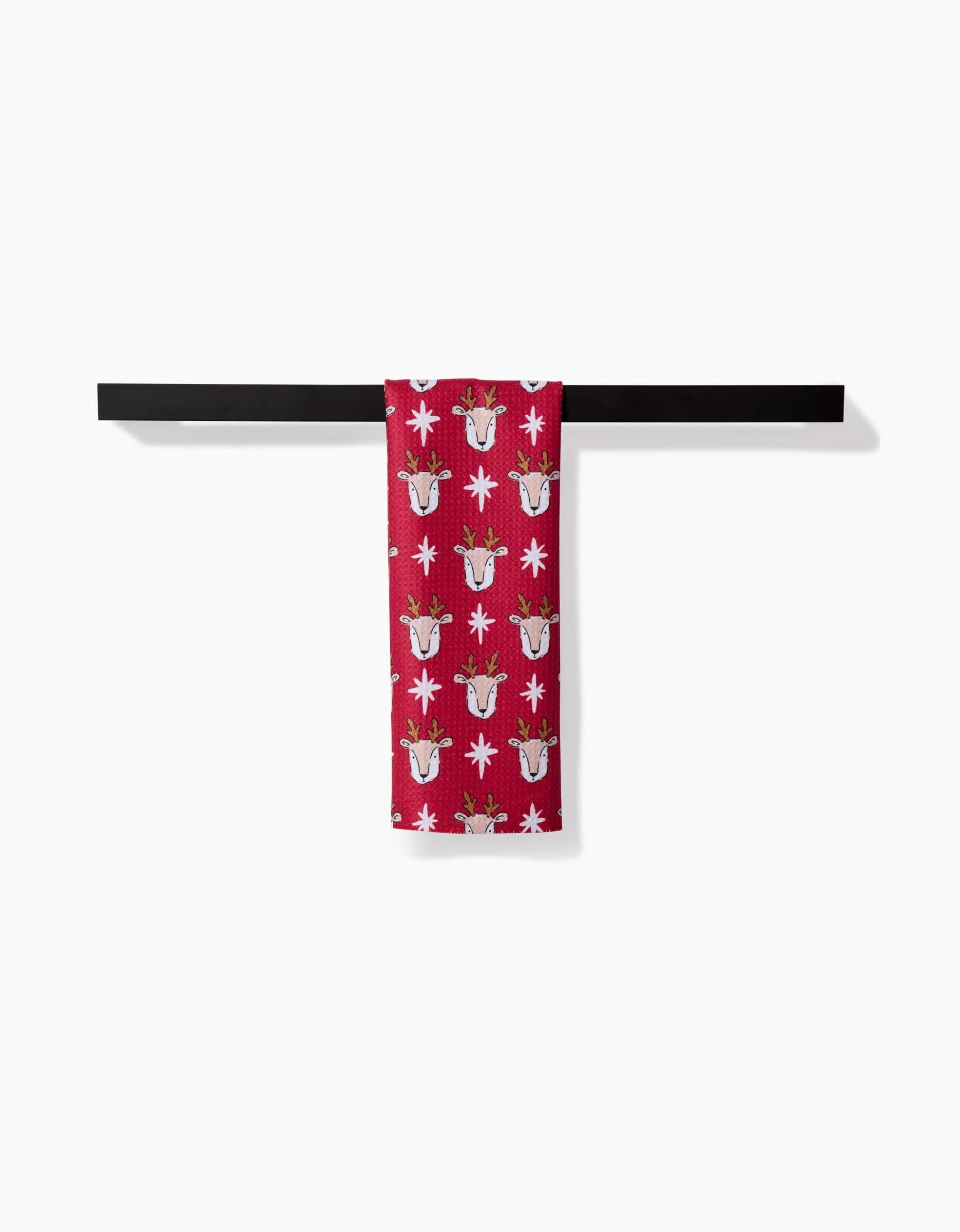 The Hufton Studio Reindeer Star bar towel, featuring festive red fabric with white and brown reindeer and snowflakes, hangs on a sleek black bar. Crafted from recycled materials, its set against a plain white background.
