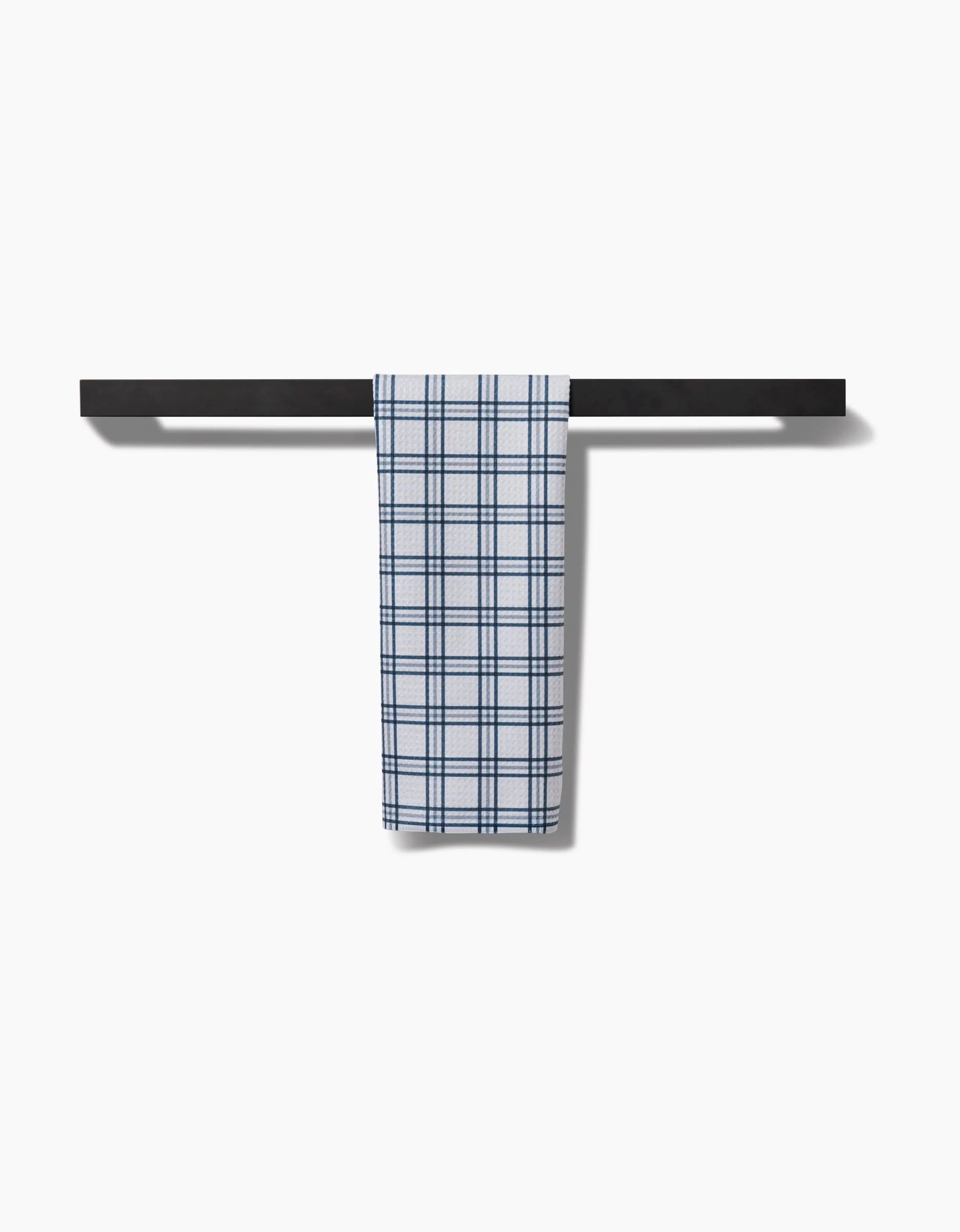 The Roma Bar Towel by Kelsi Fullmer, featuring blue and white checkered Geoweave fabric, hangs neatly on a black horizontal towel bar against a plain white background.
