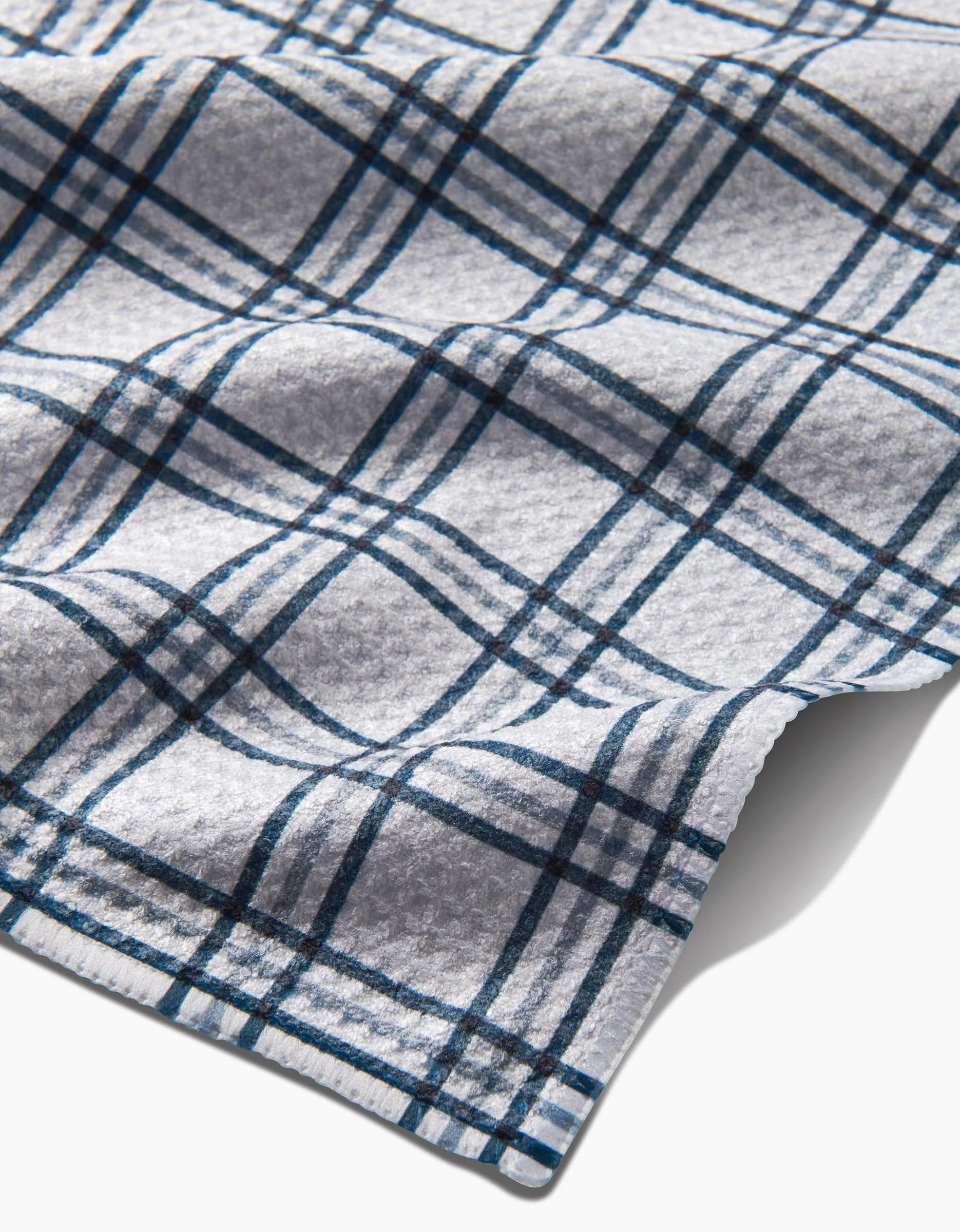 A close-up of Kelsi Fullmer’s Roma flannel shows its soft, light gray fabric with a blue and black plaid pattern, made from recycled materials. The image highlights the texture and gently curved edge of the material.
