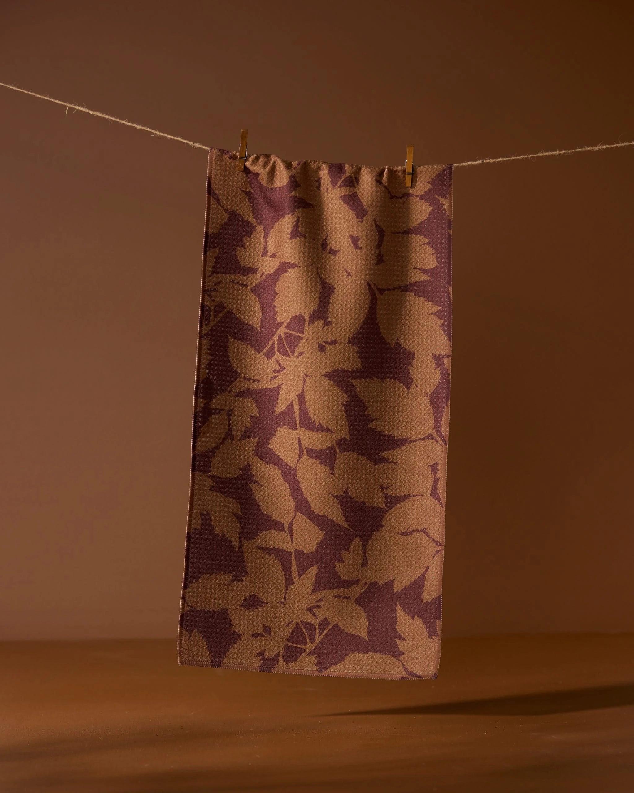 The Geometry Rustle recycled kitchen towel features a brown and tan leaf pattern, hanging on a string with two clothespins against a warm brown background.