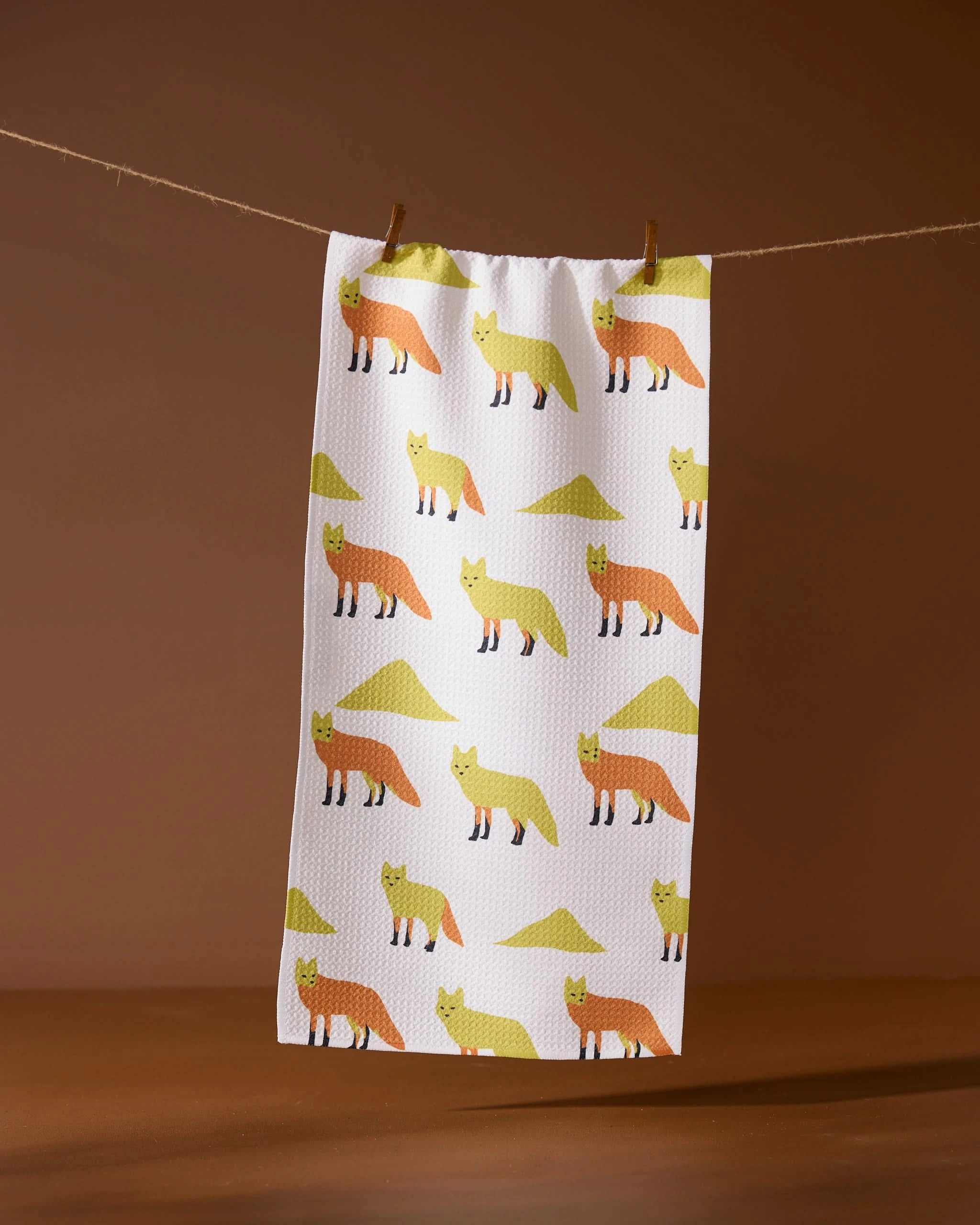 The Sagebrush Fox kitchen towel by Geometry, made from recycled materials, features orange and yellow foxes and green triangles in a repeating pattern. It hangs on a string with wooden clothespins against a brown background.