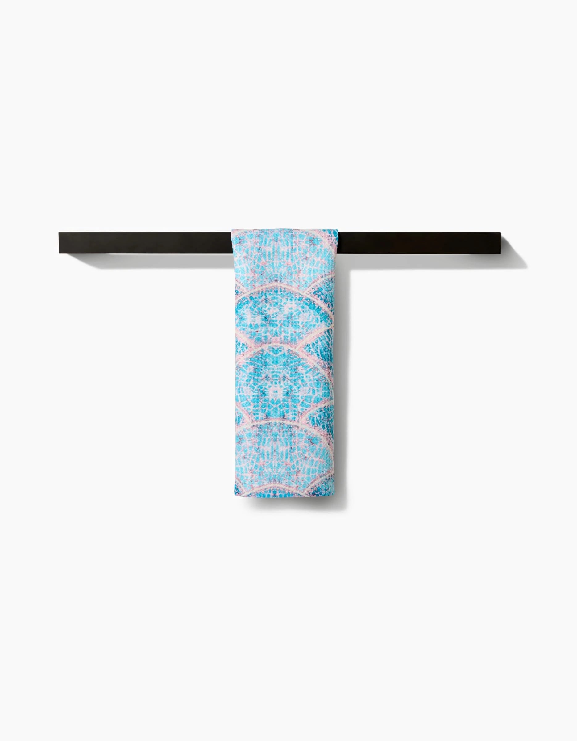 The Scallop Hills Bar Towel by Taryn Brennan, featuring a light blue and pink pattern and made from recycled materials, is draped over a sleek black towel bar against a plain white background.