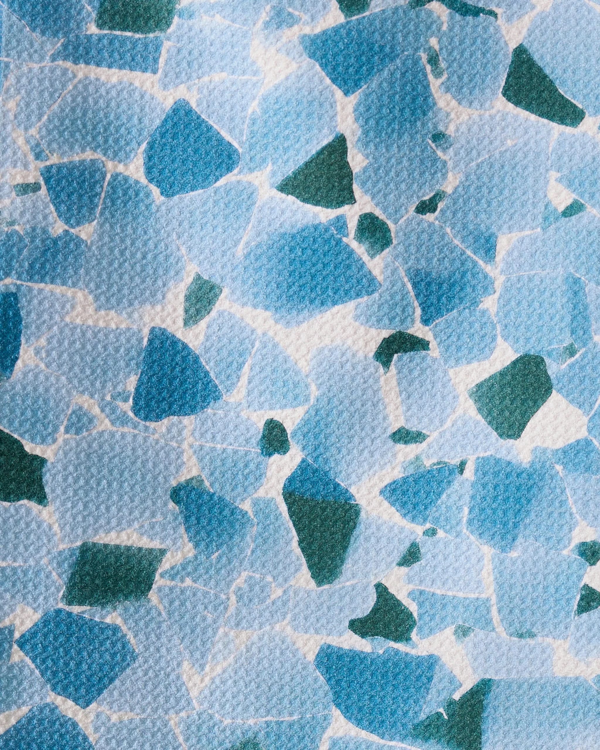 Seaglass by Geometry features an abstract mosaic of irregular blue and green shapes on a textured white background, evoking recycled materials. The random arrangement creates an artistic, modern design.