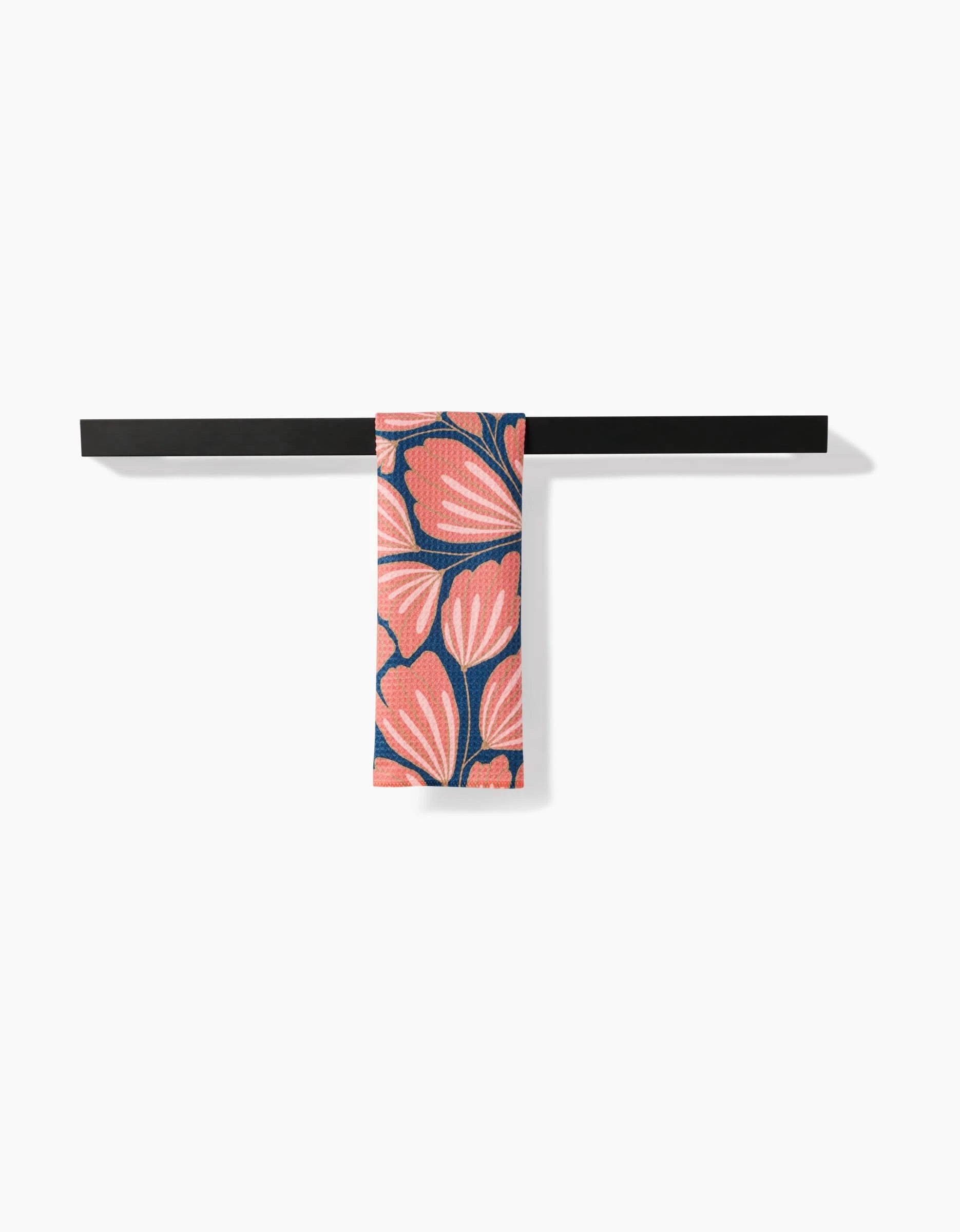 A black horizontal towel rack holds the Sheila bar towel by Amy MacCready, featuring a vibrant pink and coral floral pattern on dark blue Geoweave fabric, set against a plain white wall.