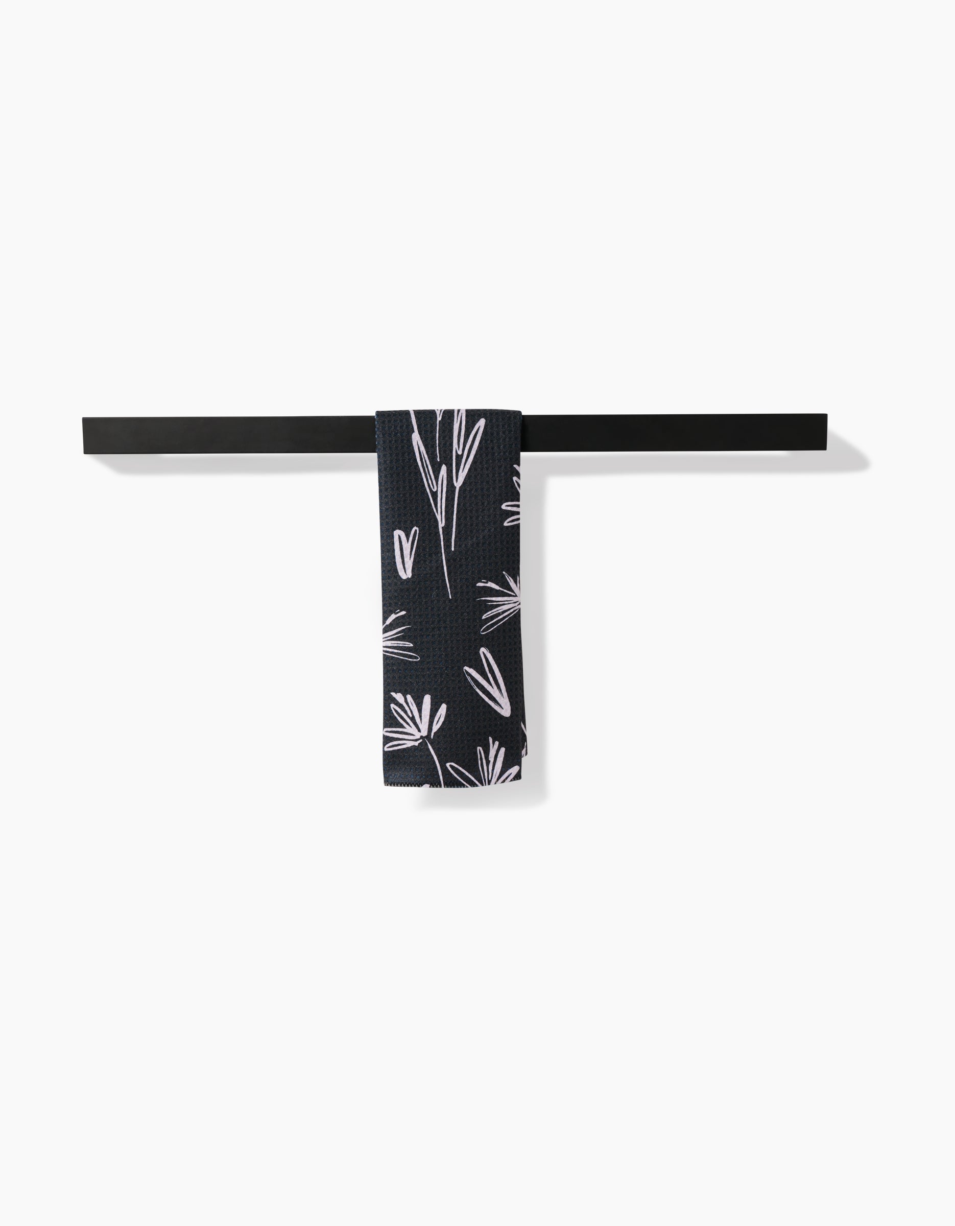 A Sketchbook Dust towel by Toboggan Avenue, crafted from soft Geoweave fabric, is neatly draped over a dark metal bar mounted on a white wall. The navy blue towel features a white floral pattern in the center.