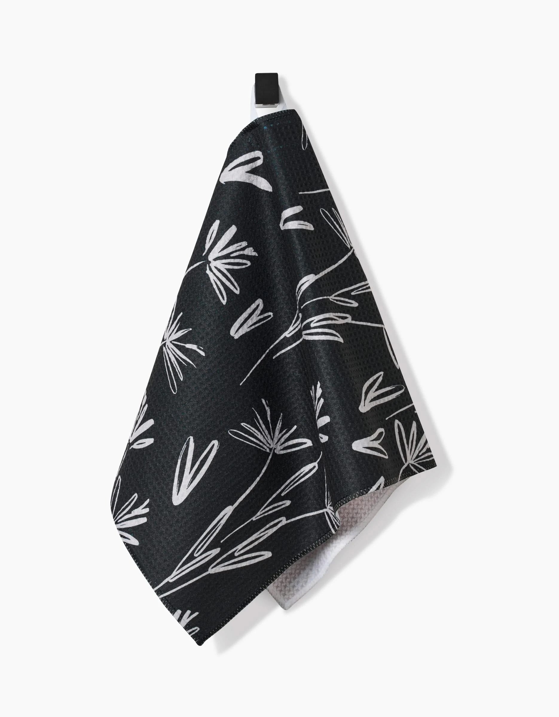 The Sketchbook Dust Bar Towel by Toboggan Avenue features a black background with a white botanical leaf and stem pattern, crafted from recycled materials, and hangs from a small black loop against a white backdrop.