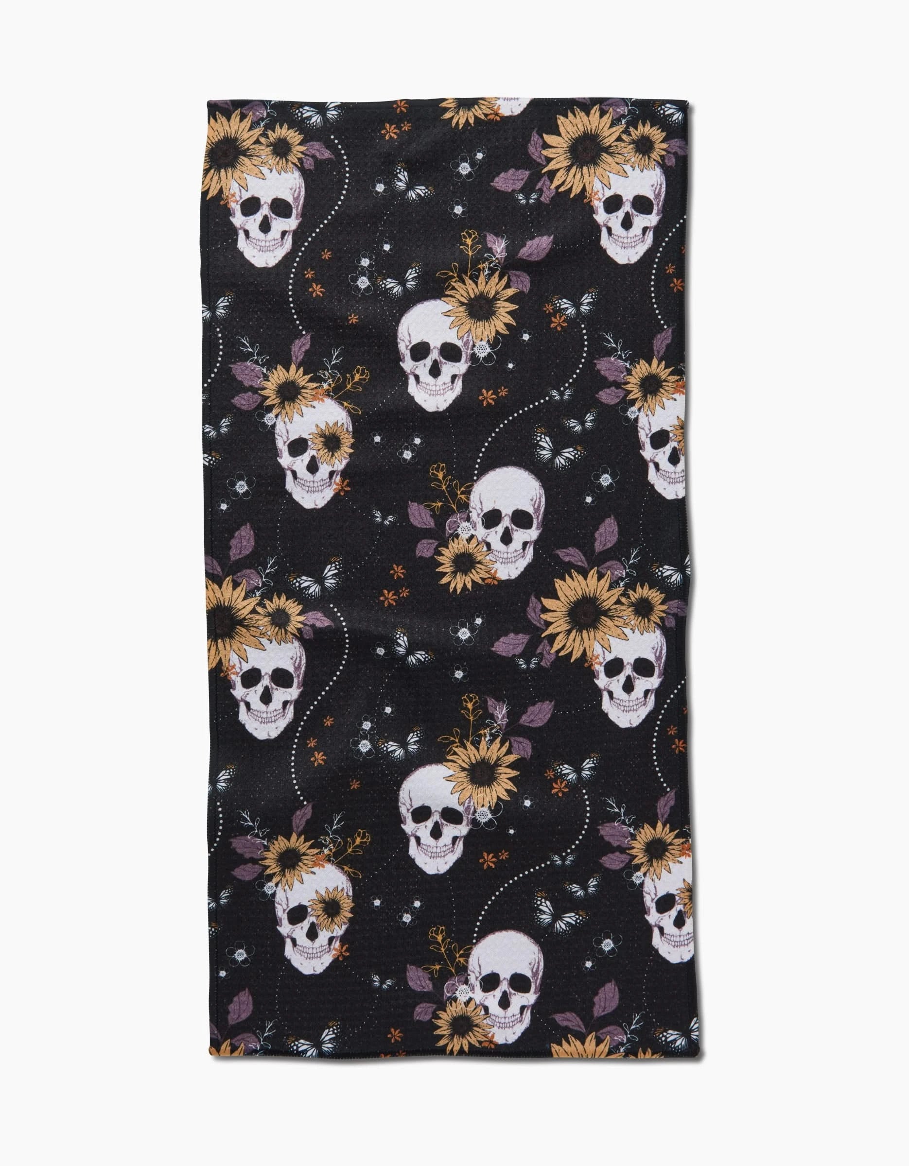 The Ashley Celena Skulls In Bloom kitchen towel is black, made from recycled fabric, and features white skulls, bright yellow sunflowers, purple leaves, and white dots in a repeating pattern.
