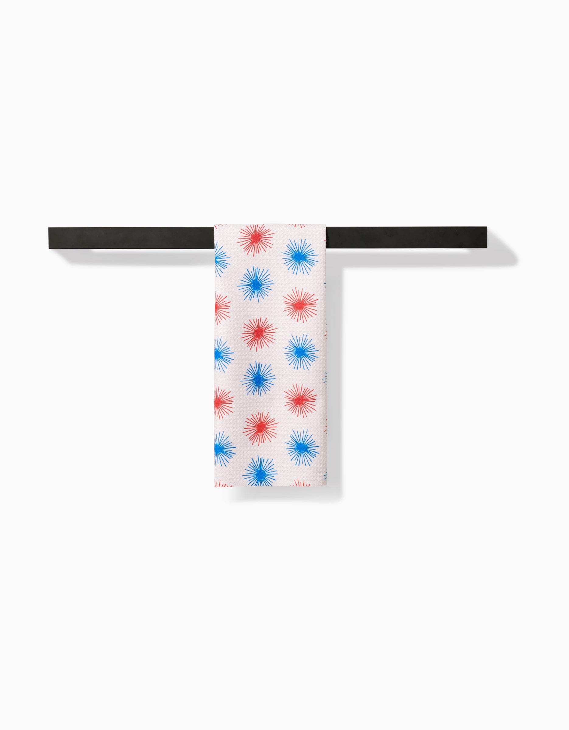 The Kayla Ann Sparkler Splash bar towel, crafted from Geoweave fabric with red and blue starburst patterns, hangs neatly over a black rod against a plain white background.