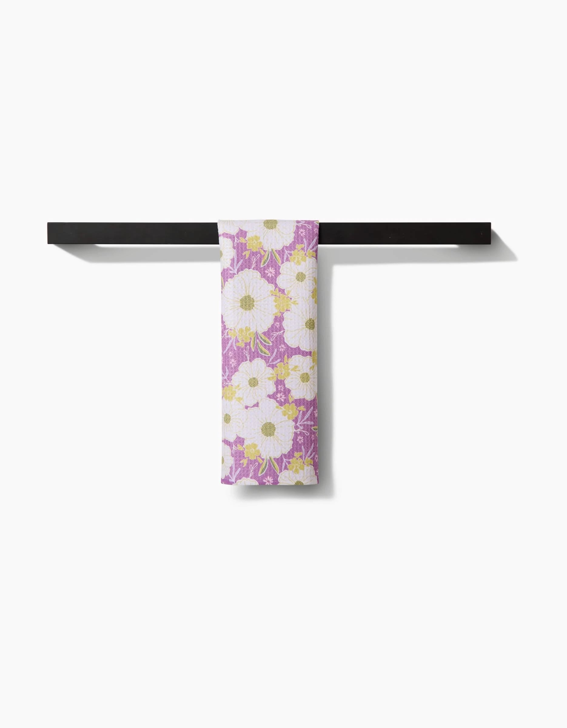 The Ashley Celena Springtime Floral bar towel, featuring white and yellow daisies on a purple background, hangs neatly folded over a black towel bar against a white backdrop. Crafted from recycled materials.