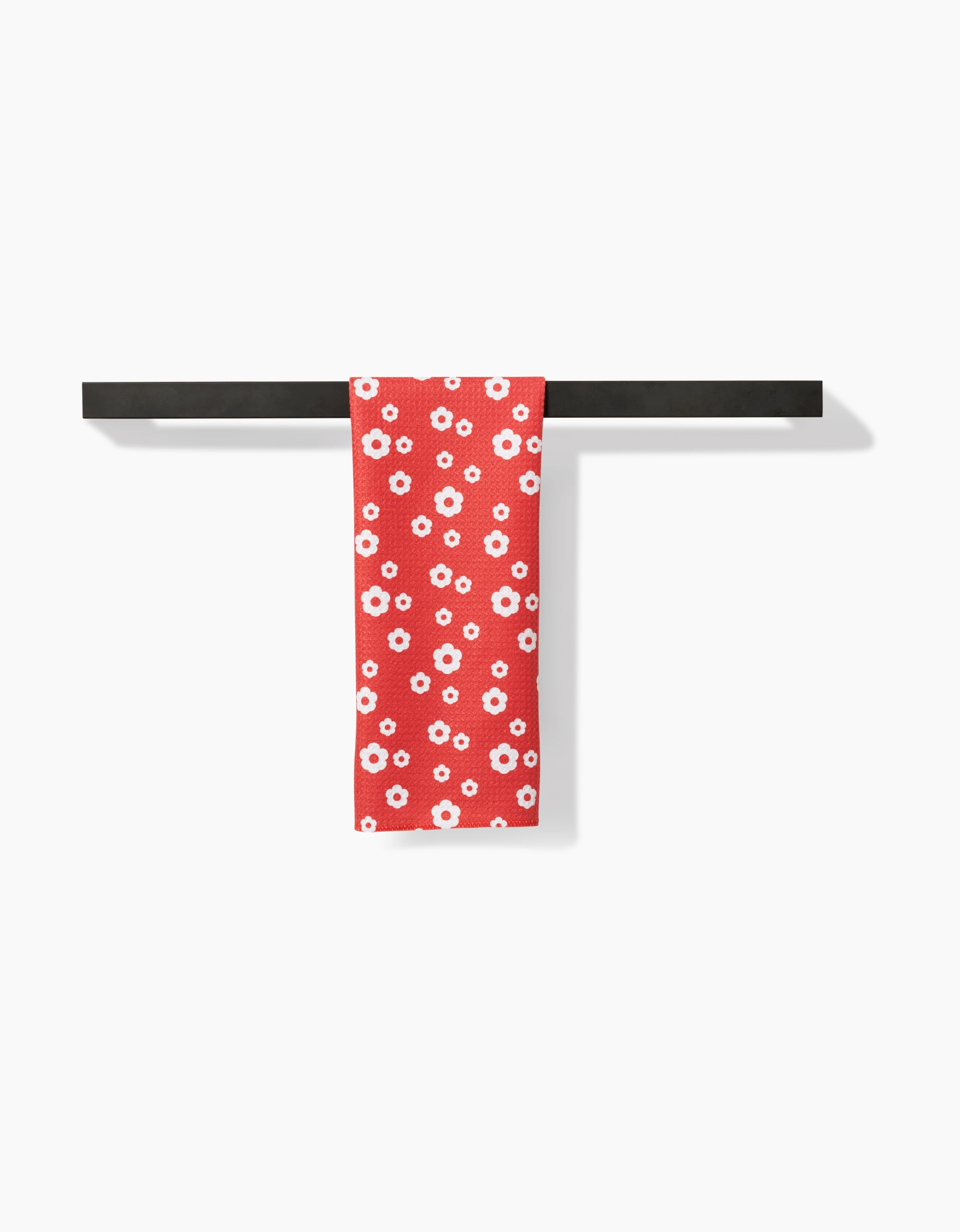 The Sunny Flowers towel by Danelys Sidron, featuring a white floral pattern on red Geoweave fabric, hangs neatly over a horizontal black bar towel rack against a plain white background.