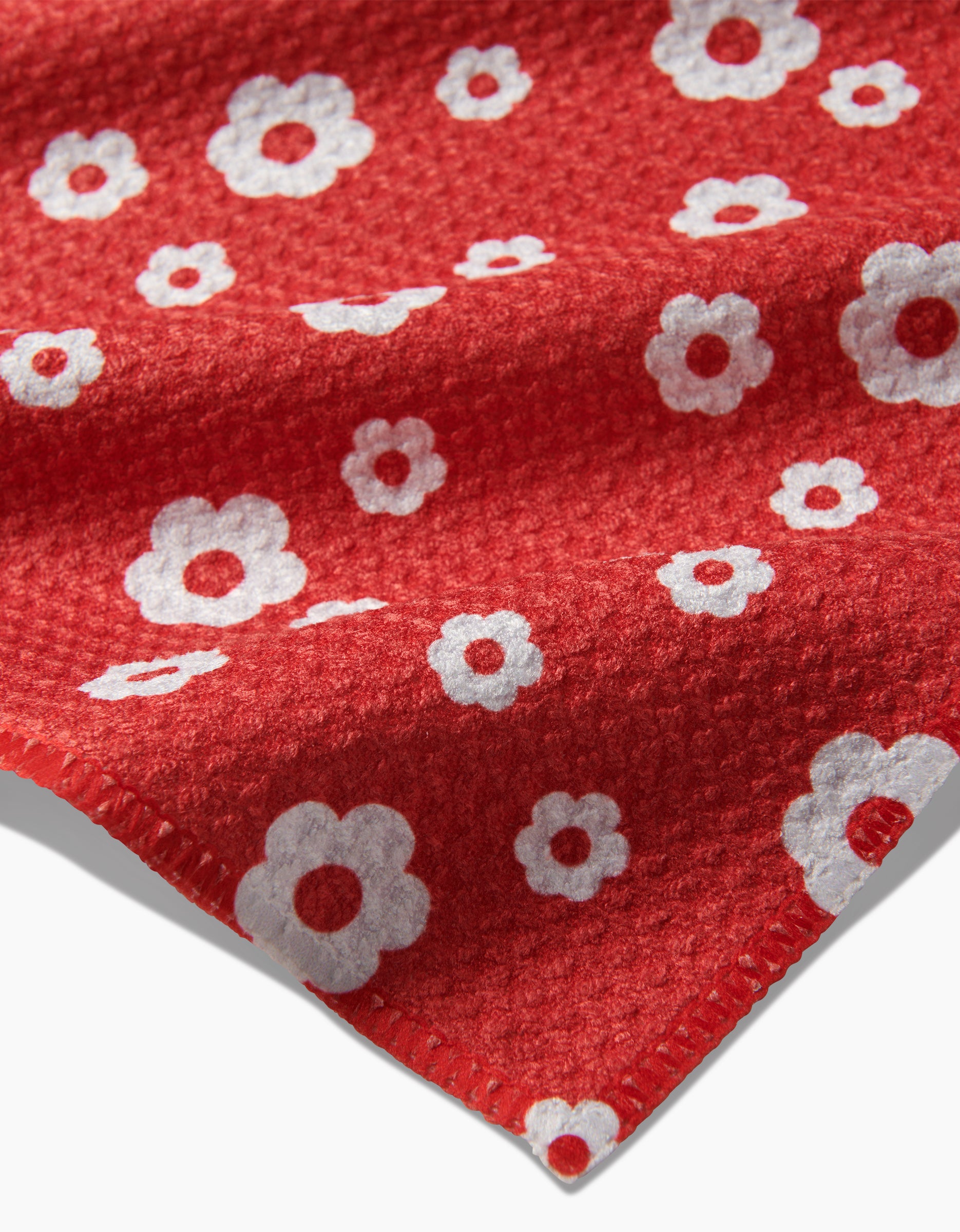 A close-up of Danelys Sidron’s Sunny Flowers bar towel shows its red textured Geoweave fabric, adorned with white flowers in assorted sizes and a visible edge in the foreground.