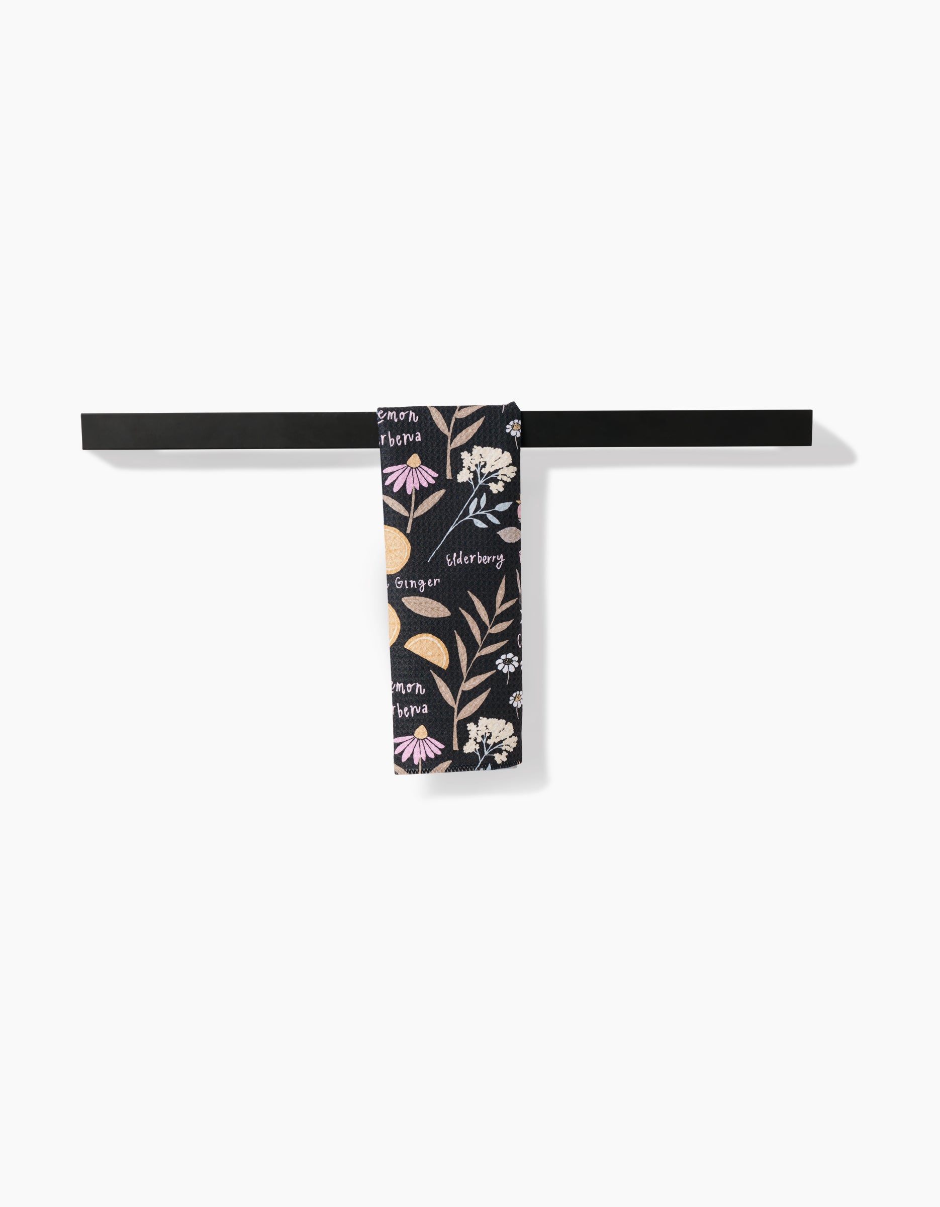 A sleek black metal bar towel holds the Sweet Tea towel by Janna Sue Design, featuring labeled herbs and flowers in a botanical print on a dark background.