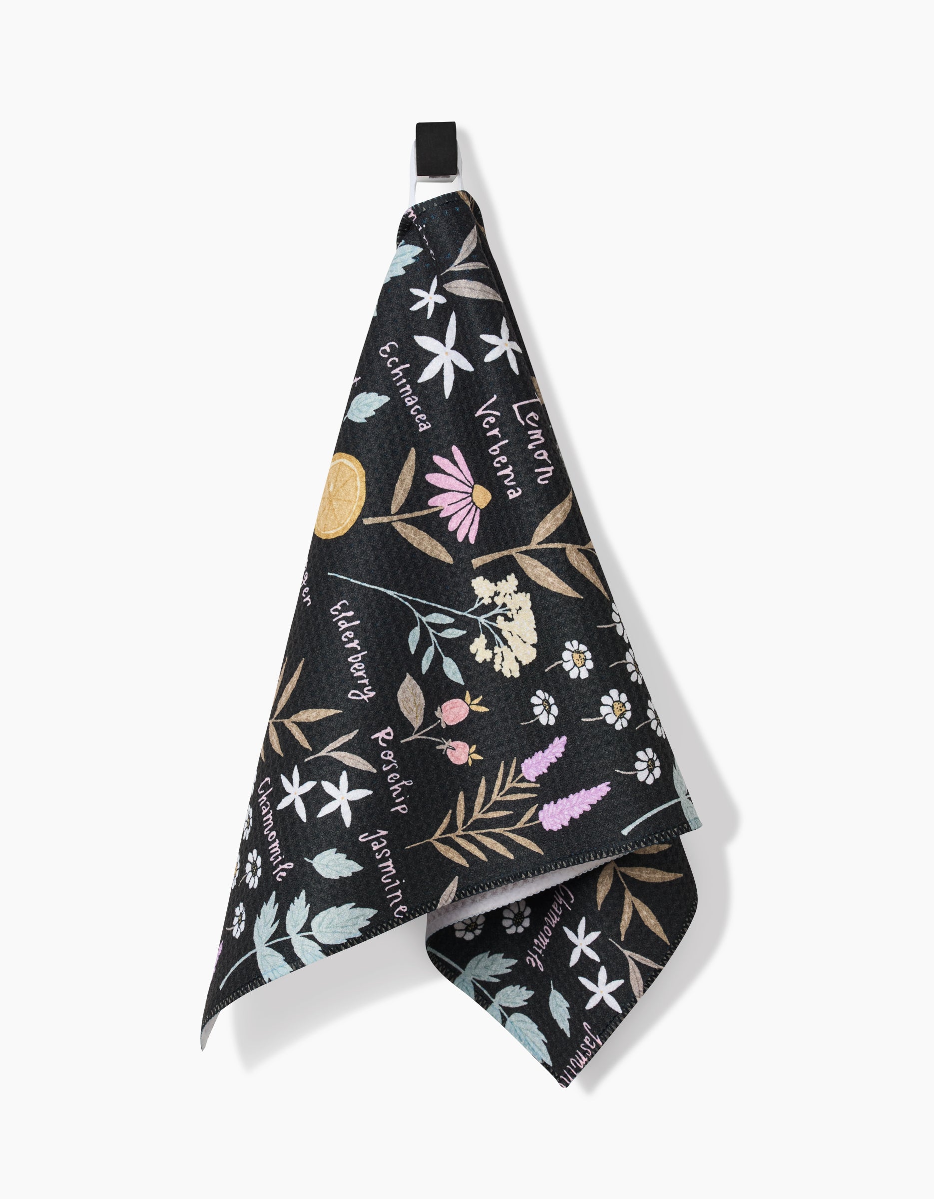 A black Sweet Tea bar towel by Janna Sue Design hangs on a hook, adorned with vibrant floral art and cursive herb names like echinacea, lavender, and rosemary. Eco-friendly and crafted from recycled materials.