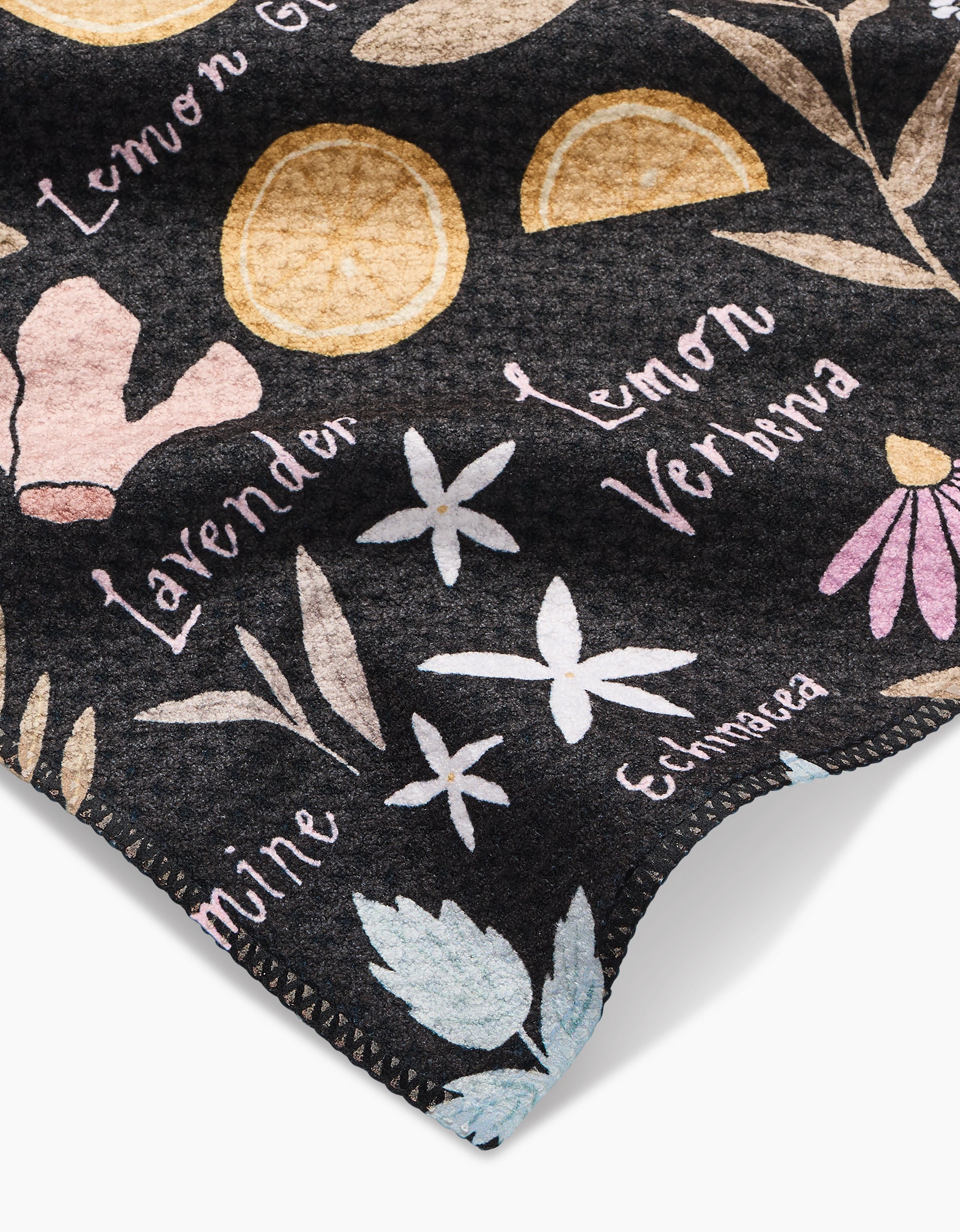 The Sweet Tea Bar Towel by Janna Sue Design features dark Geoweave fabric with vibrant botanical illustrations and labeled herbs like lemon, ginger, lavender, lemon verbena, echinacea, and mint. Stitched edges add durability.
