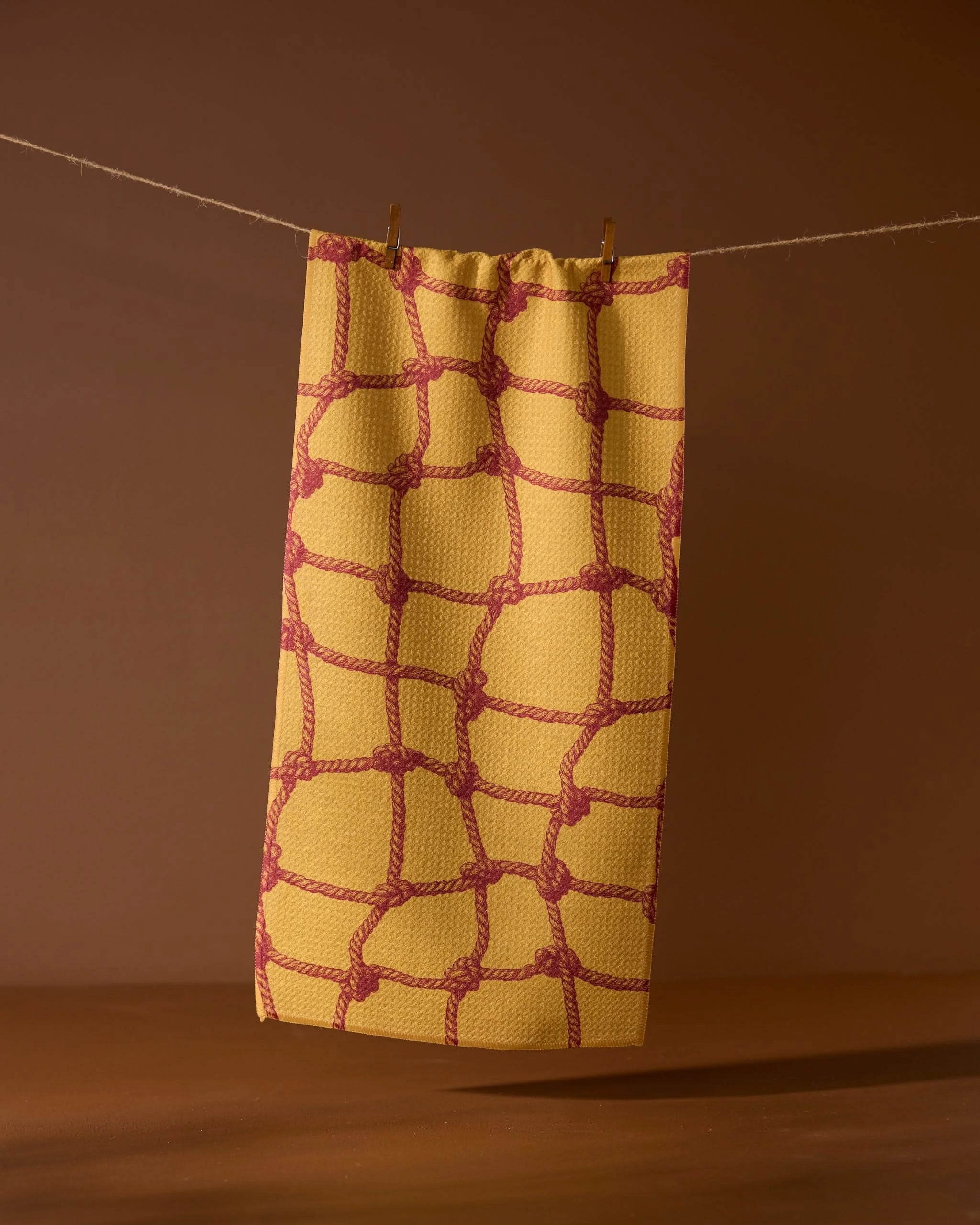 The Tethered Orange kitchen towel by Geometry, featuring a red rope knot pattern, hangs from a string with clothespins against a brown background.