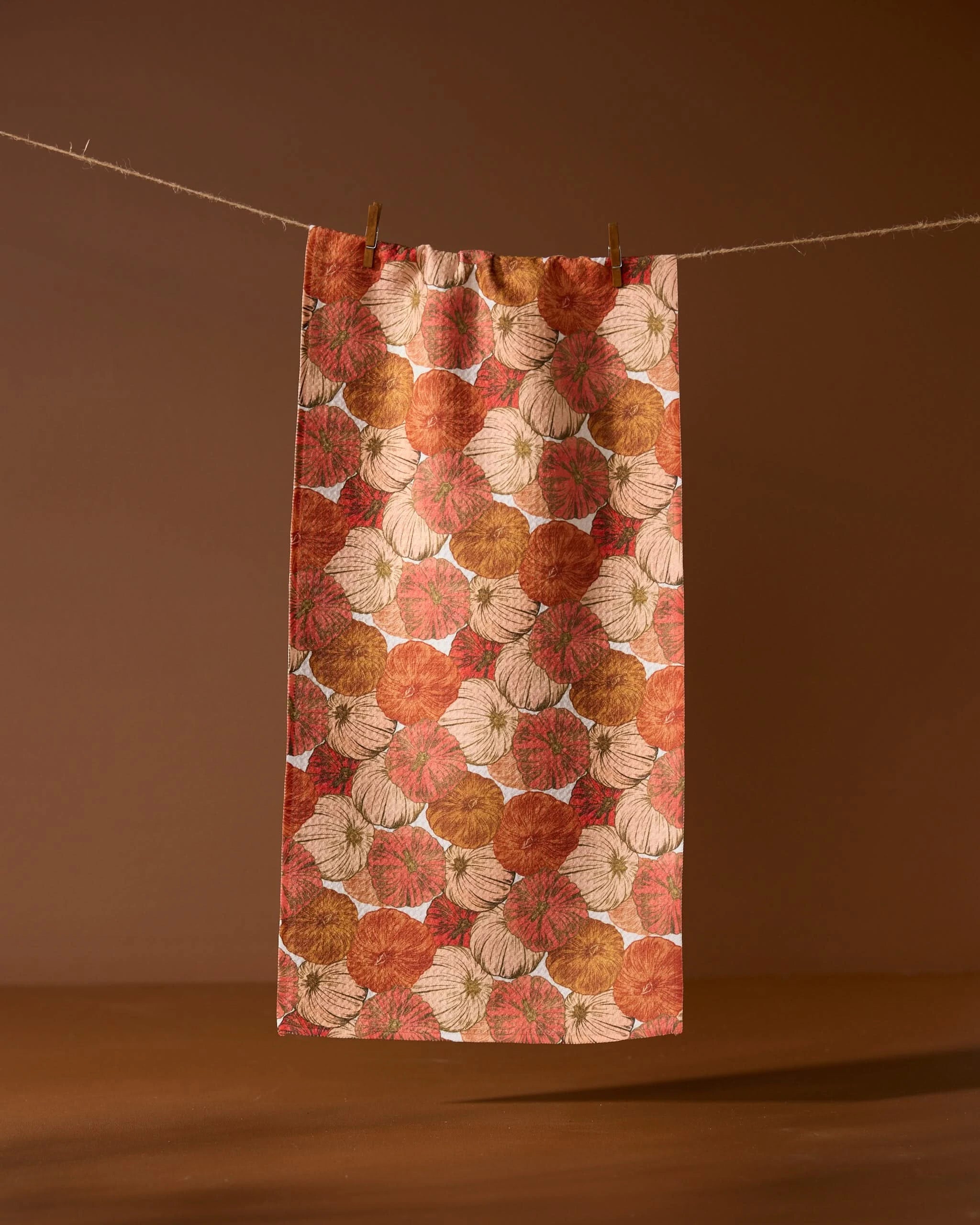 The Geometry Tendril towel, featuring an ultra-absorbent design with orange, brown, and beige pumpkin patterns, hangs on a string against a plain brown background, held by two wooden clothespins.