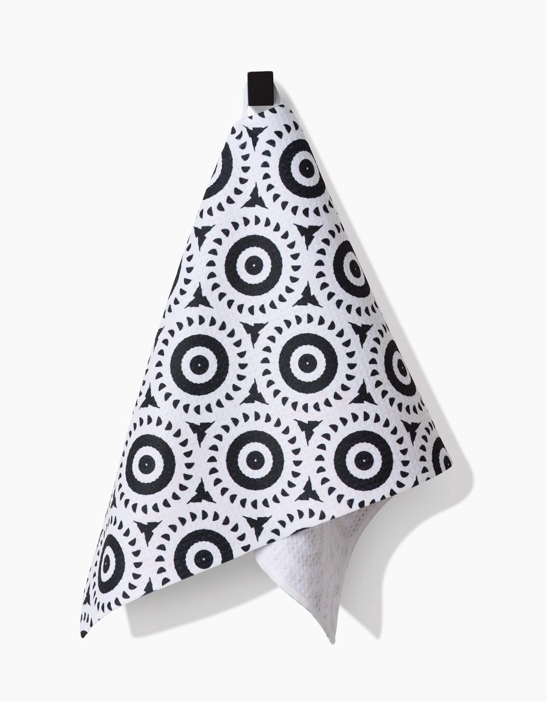 The Texas Two Step bar towel by Katherine Berkeley hangs from a black hook, showcasing bold black concentric rings with triangle details. Made of durable Geoweave fabric, the white towel casts a shadow on a white background.