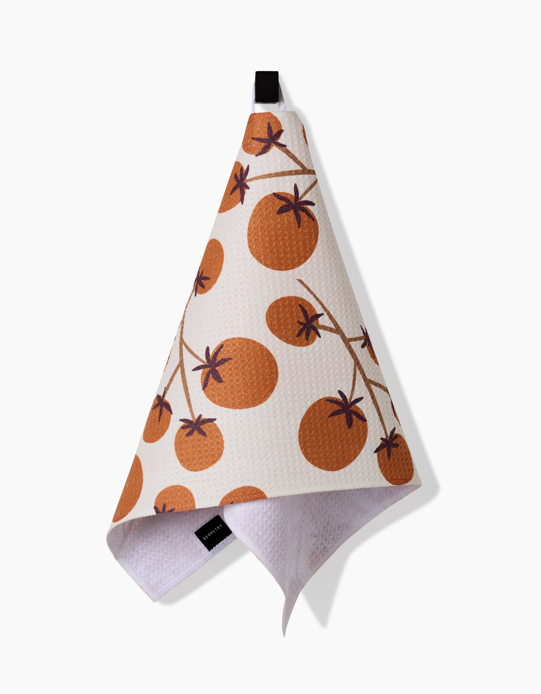 The Lizzie Clark Tomato Harvest bar towel, made from recycled materials and featuring a white textured fabric with an orange tomato pattern, hangs from a black loop against a plain white background.