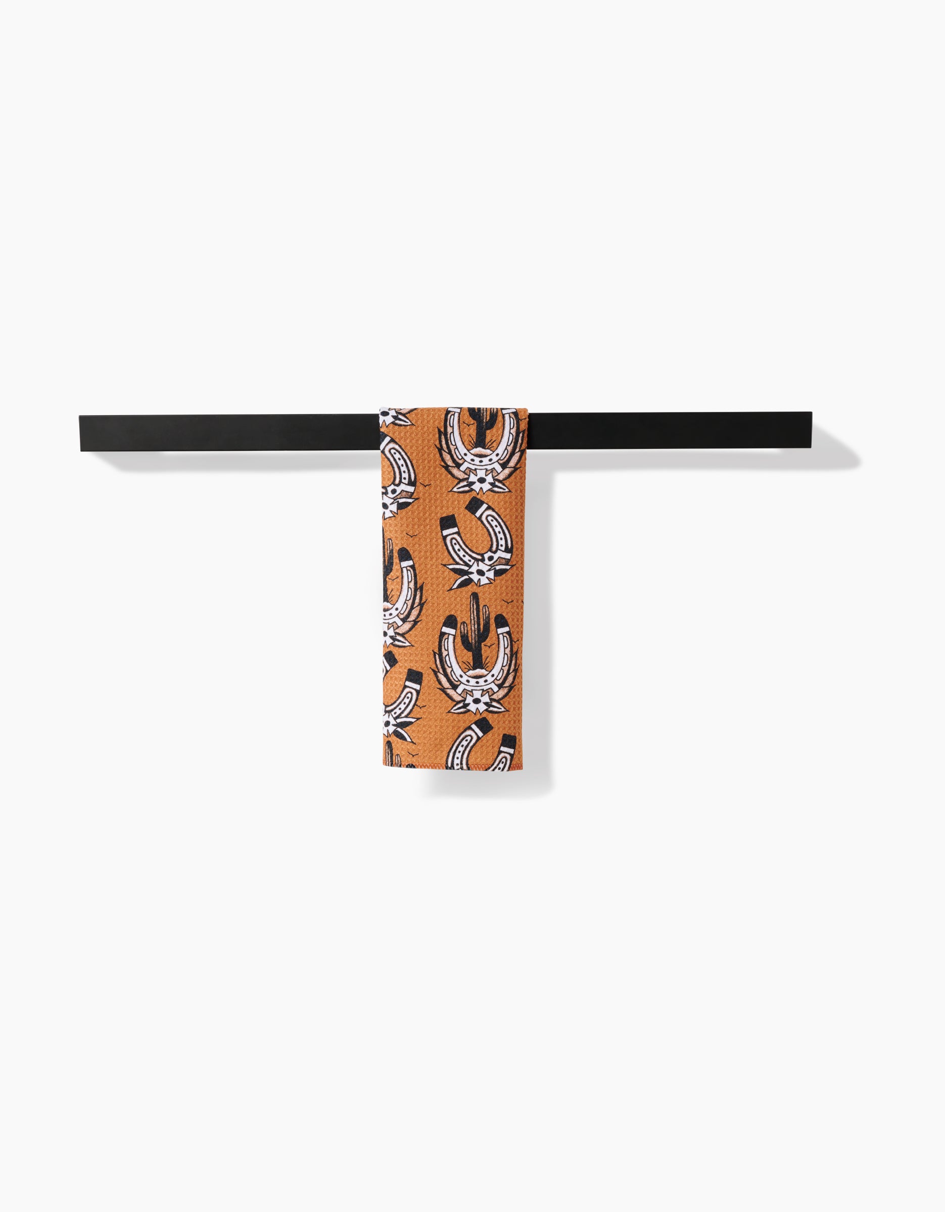The Cole Strem Western Horseshoes bar towel, featuring white and black patterns on orange recycled fabric, hangs neatly on a black towel bar against a plain white background.