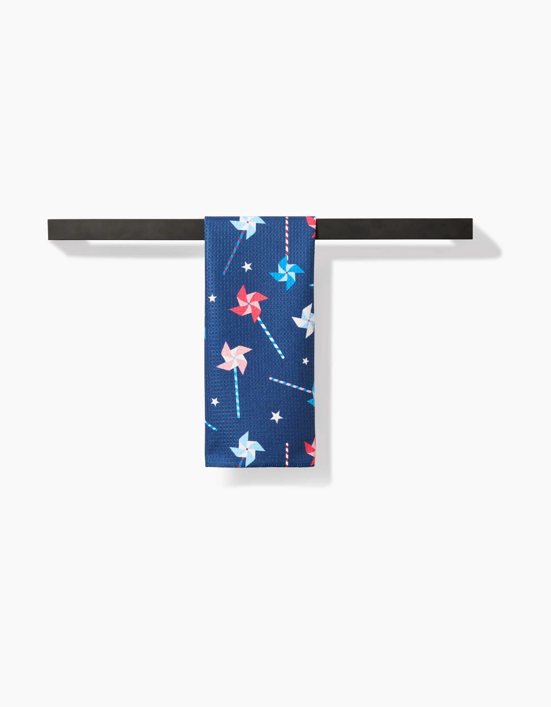 The Rebecca Bobko Whirlwind Pins bar towel, featuring red, white, and blue pinwheels with white stars on navy Geoweave fabric, hangs neatly on a black rod against a white background.