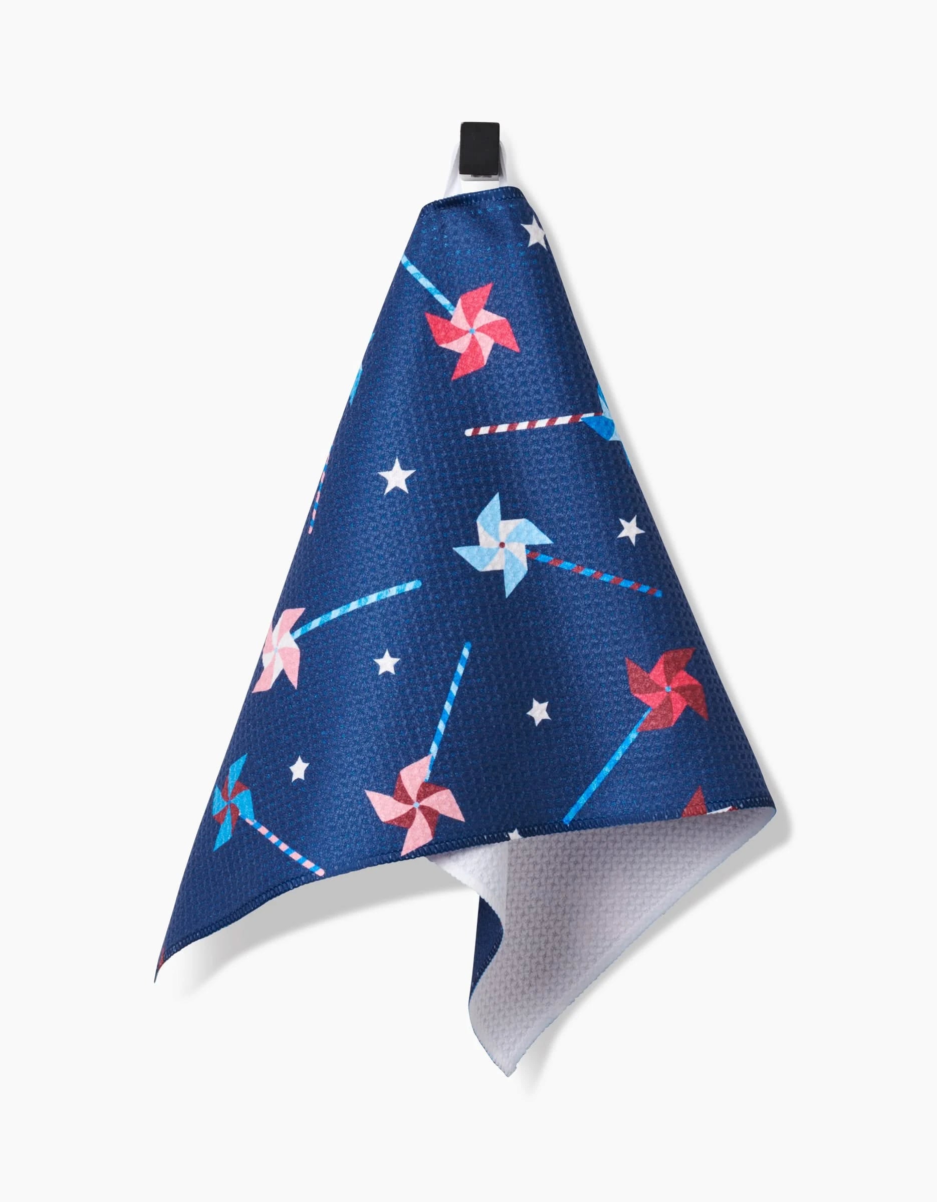 The Whirlwind Pins bar towel by Rebecca Bobko is made with Geoweave fabric and features a pattern of red, white, and blue pinwheels with white stars. It hangs by a black loop against a white background.