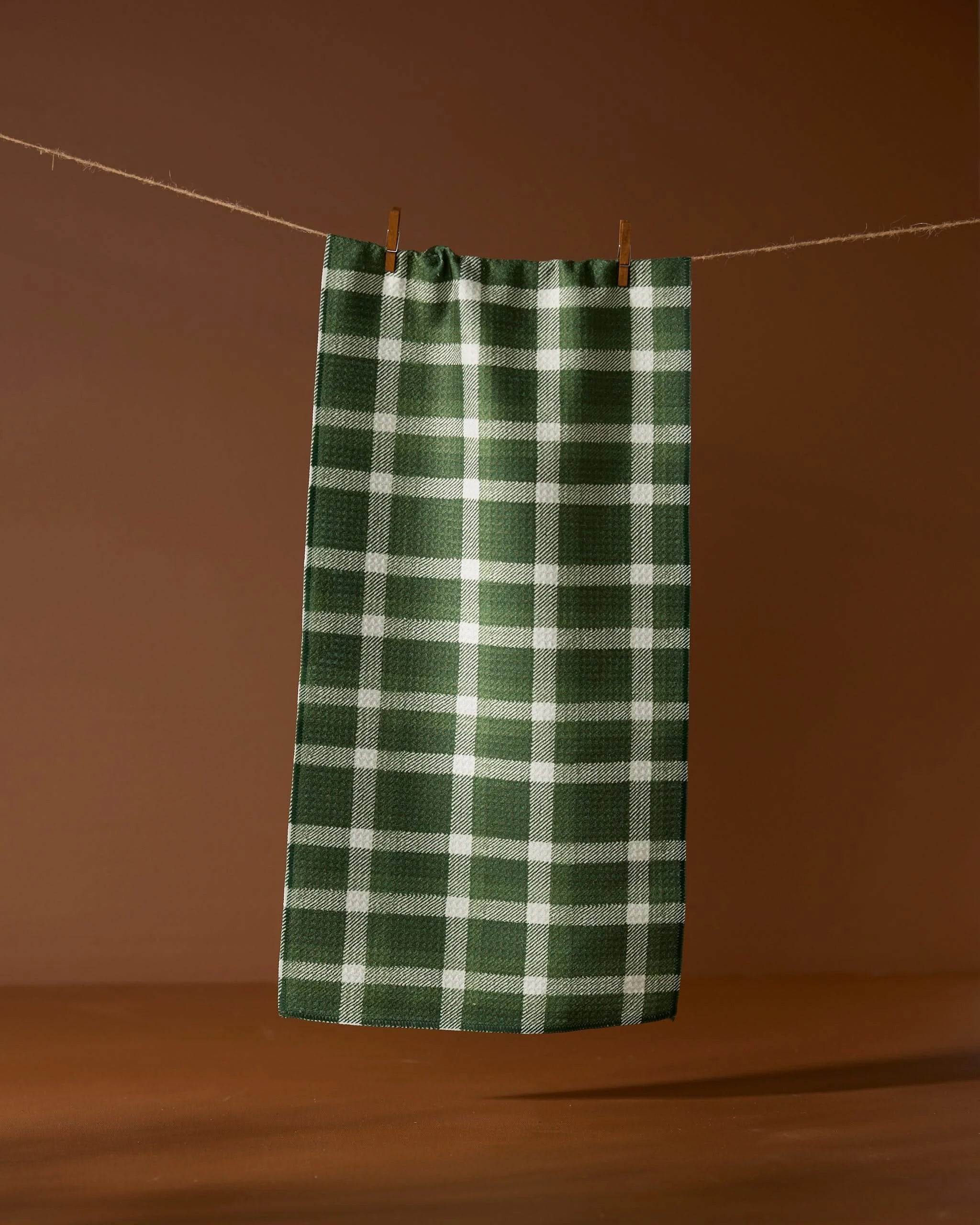 The Geometry Wicklow Green ultra-absorbent kitchen towel, featuring green and white checks, hangs on a clothesline against a brown background.