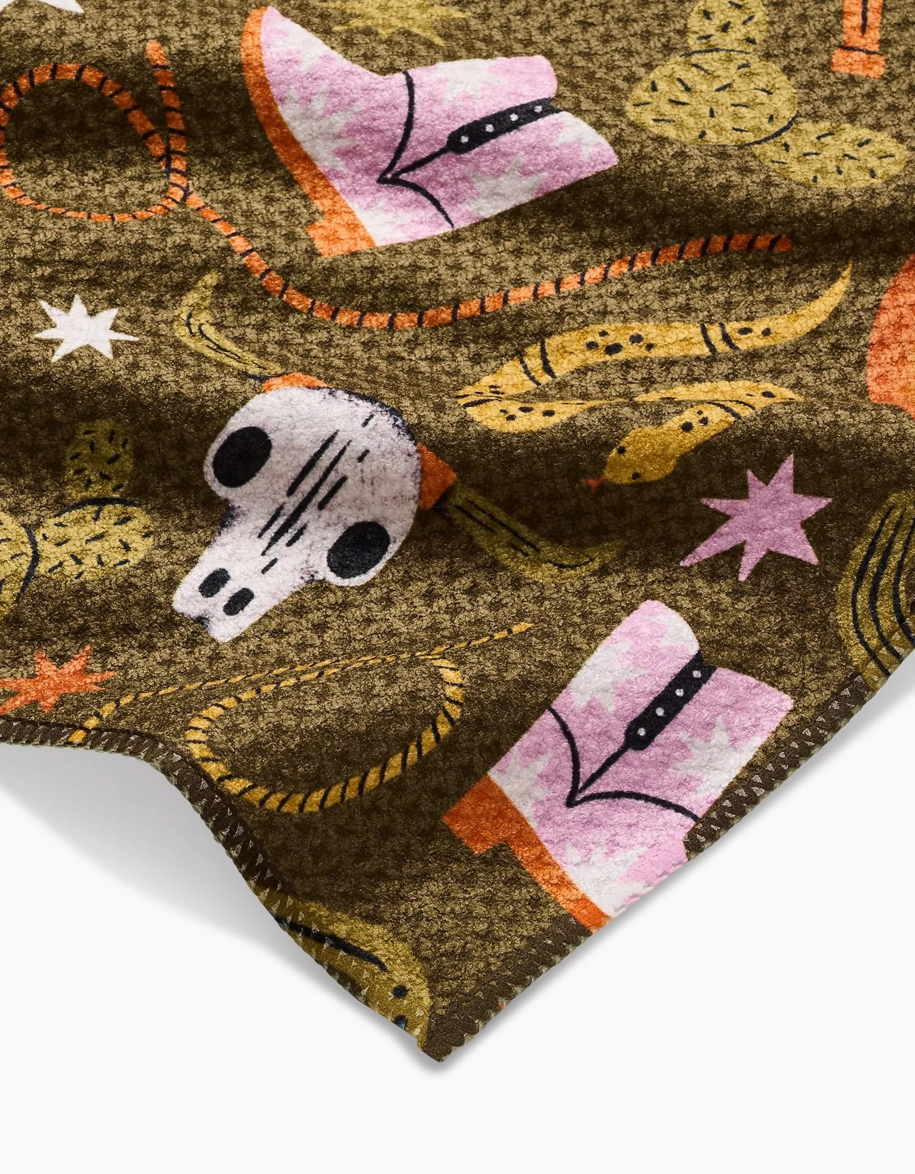 A close-up of the Megan Roy Wild Cowboy Bar Towel, featuring pink cowboy boots, a white cow skull, yellow cacti, lasso ropes, and stars on brown Geoweave fabric with visible edge stitching.